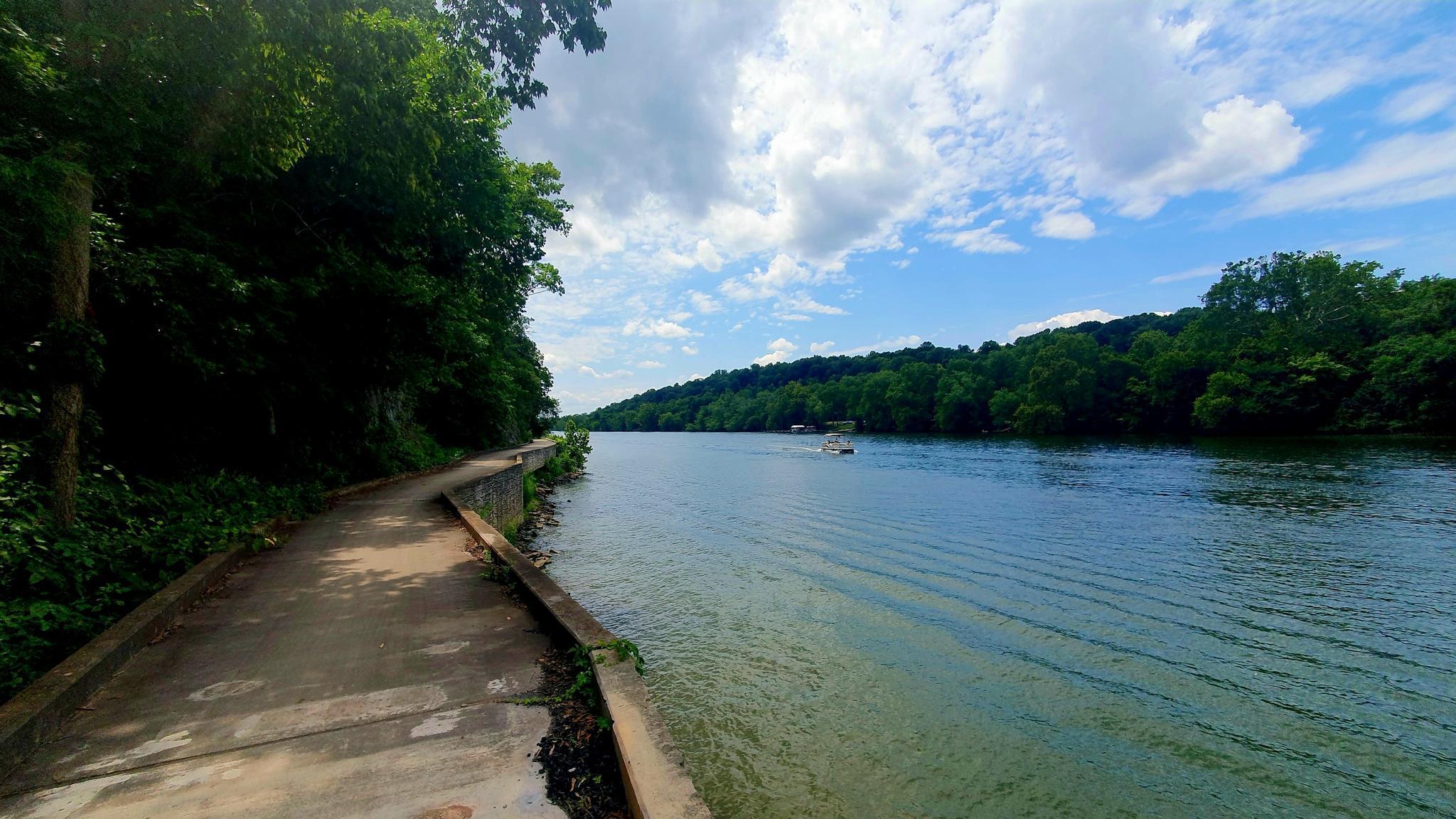 Concrete path next to a wide river with a distant boat, partly cloudy.