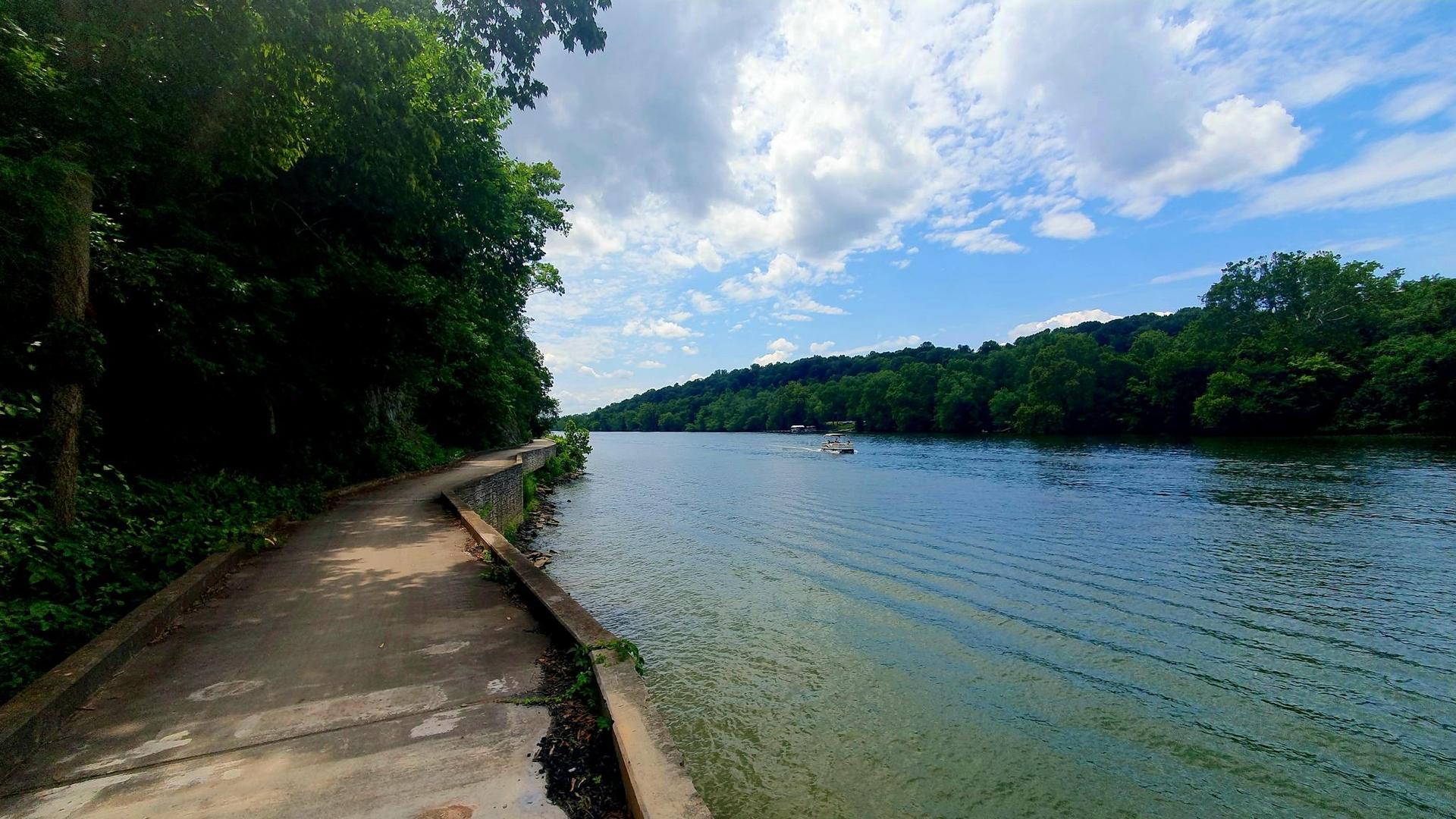 Concrete path next to a wide river with a distant boat, partly cloudy.