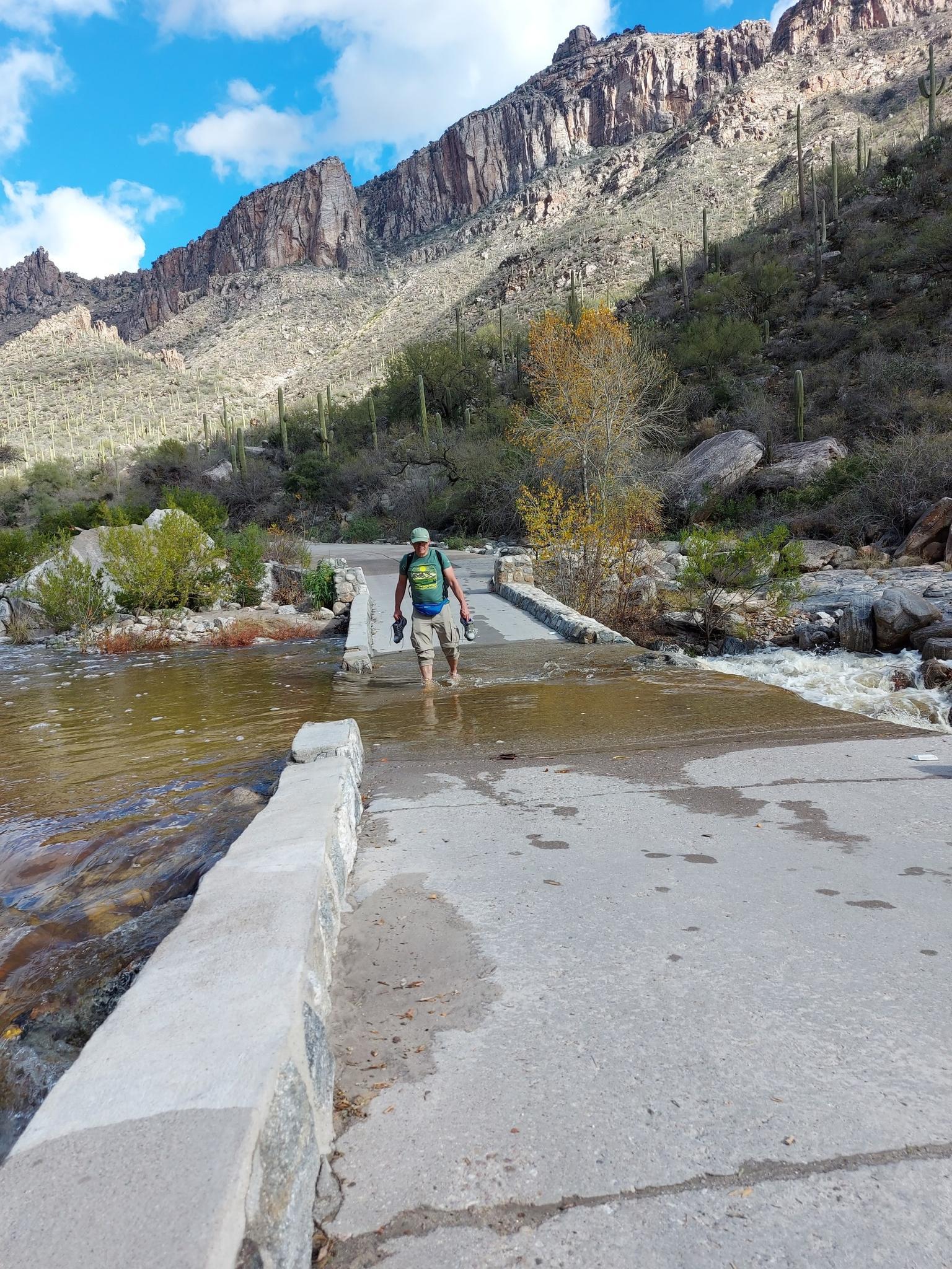 Man with backpack walks through shallow river on a concrete crossing.