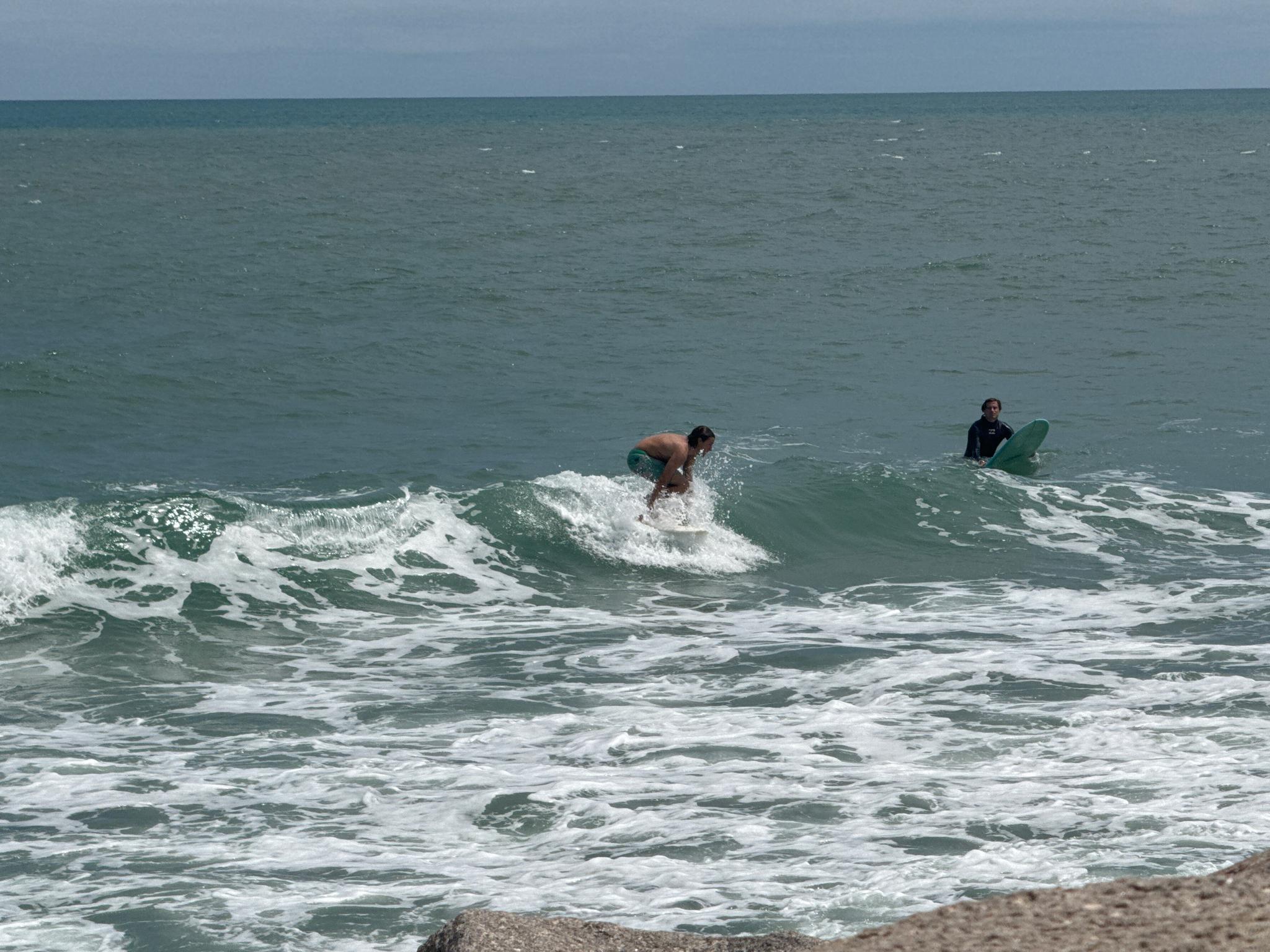 Two people in the ocean, one surfing a small wave under a bright sky.
