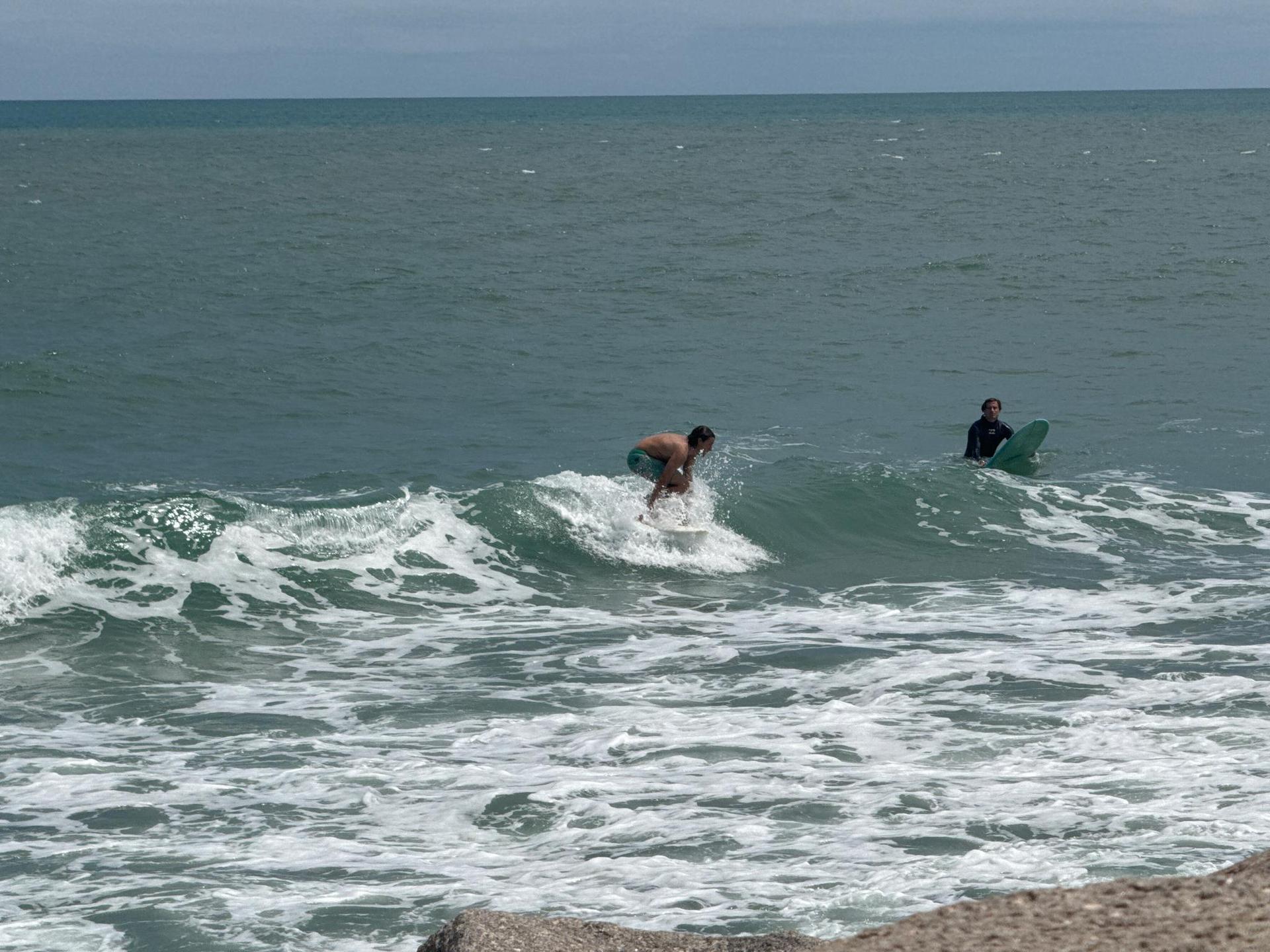 Two people in the ocean, one surfing a small wave under a bright sky.