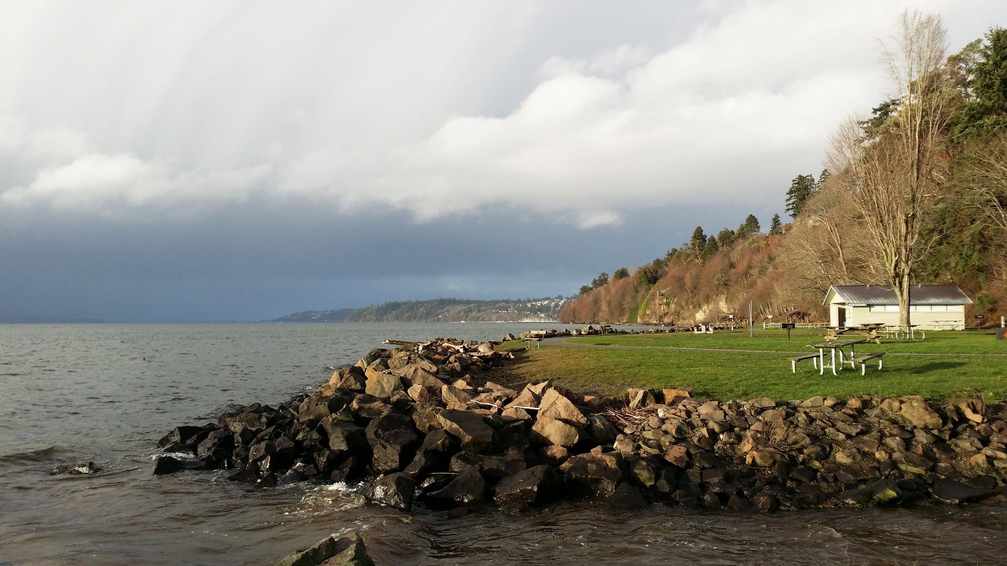 Rocky shoreline with a small building, green grass, and trees under a cloudy sky. - North and South Rim Trail Loop