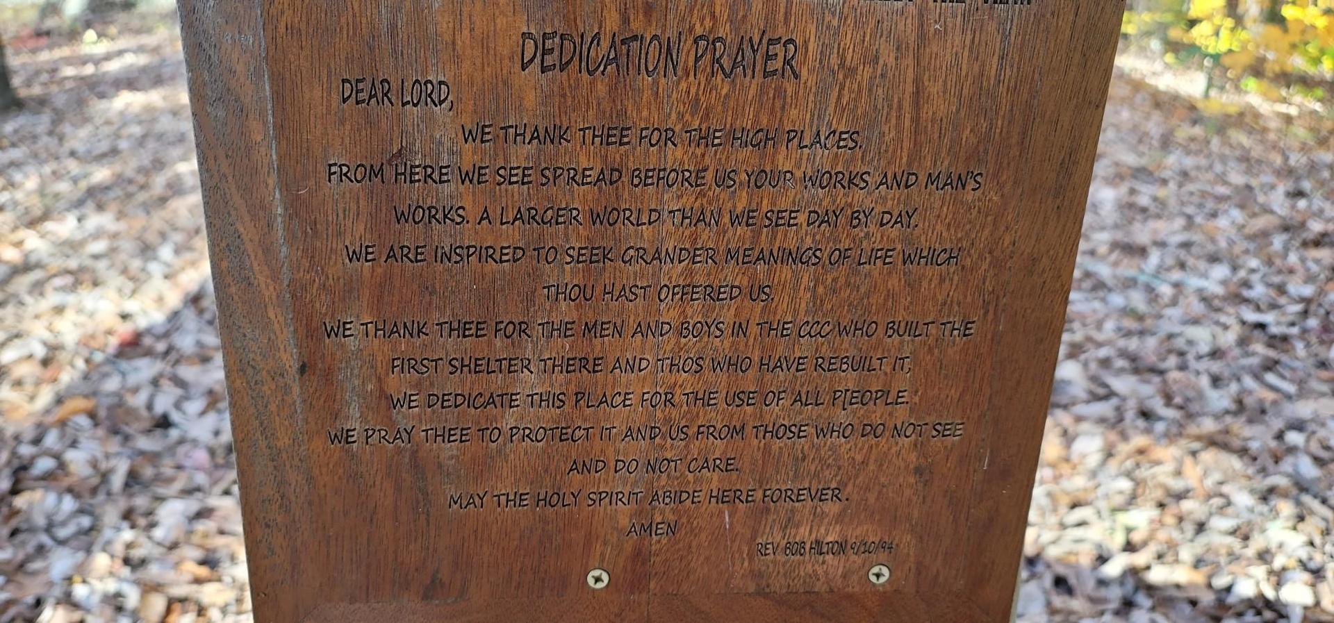 A wooden plaque with an engraved dedication prayer on a forest floor.