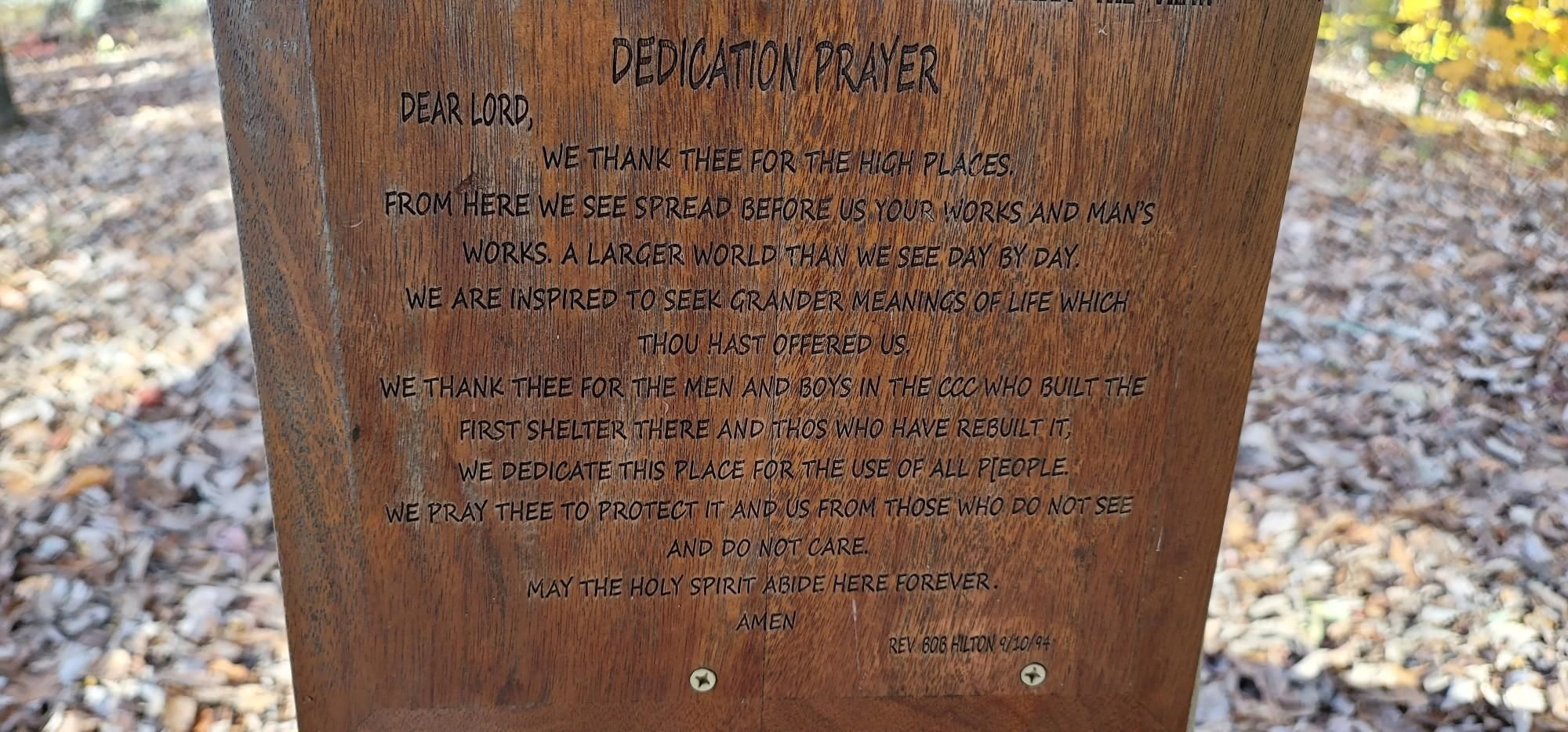 A wooden plaque with an engraved dedication prayer on a forest floor. - Observation Point Trail