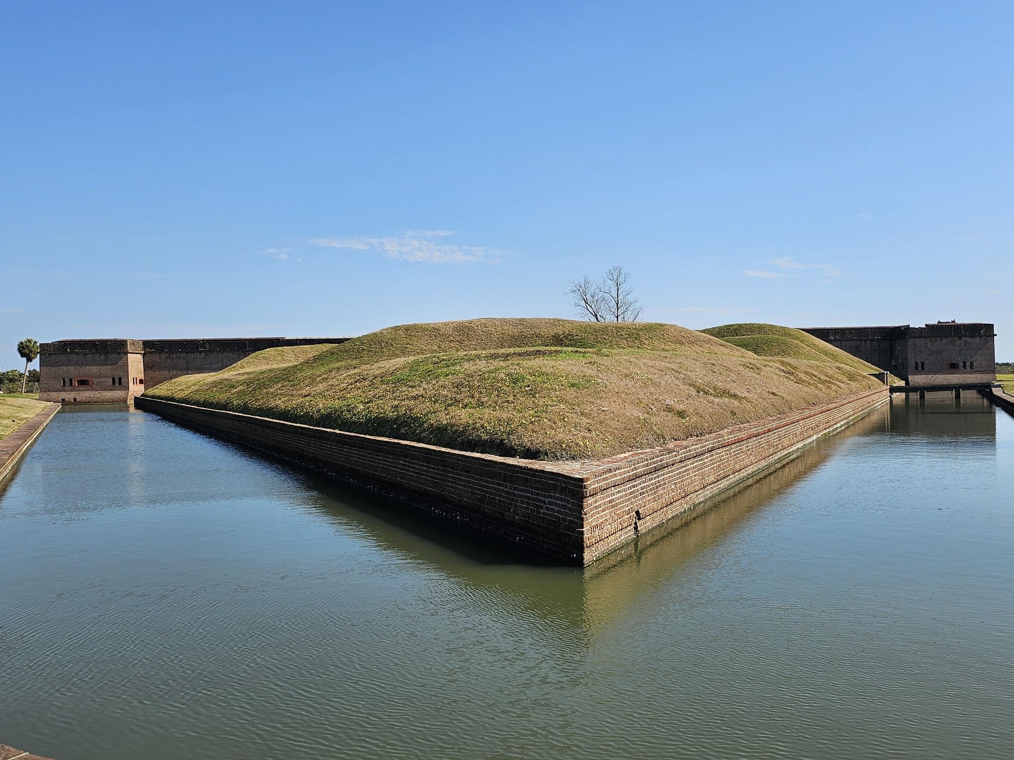 Grassy triangular mound surrounded by water, with stone walls under blue sky.