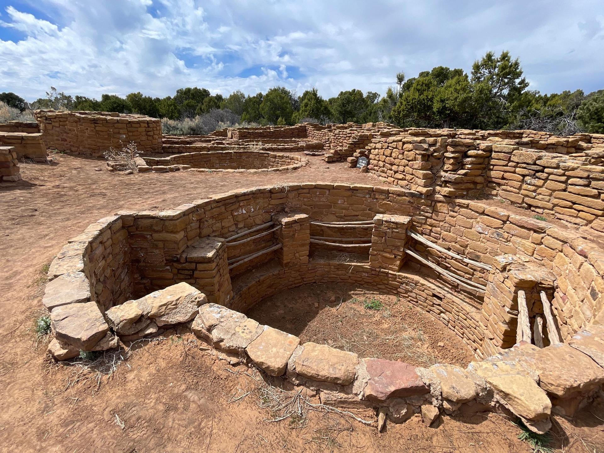 Circular stone pit with benches, surrounded by other ruins under a cloudy sky.