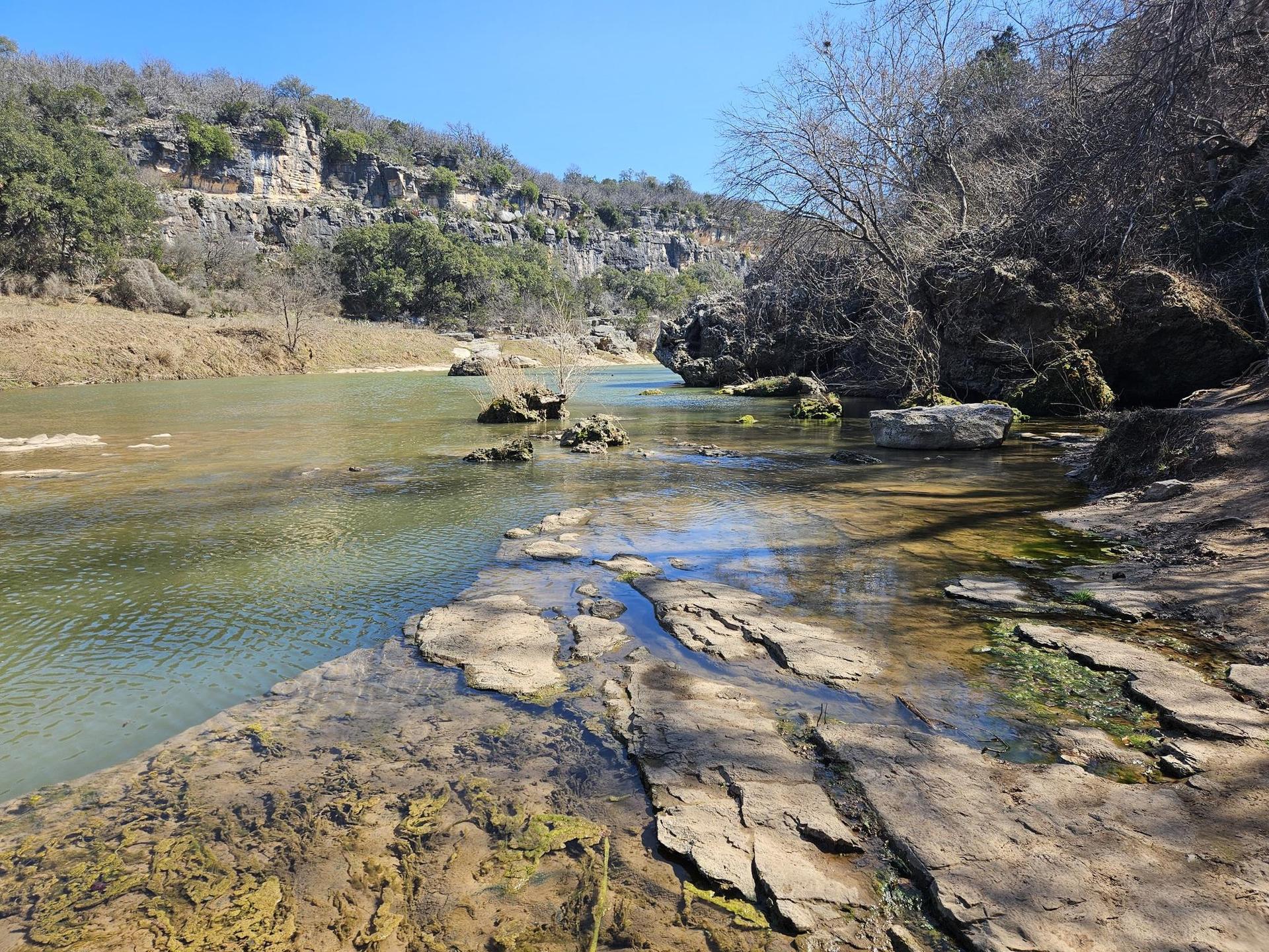 Clear river flowing over rocky beds, surrounded by cliffs and trees.