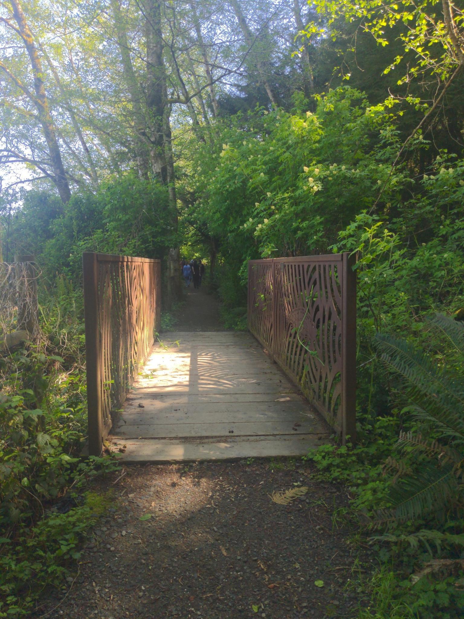 Wooden bridge with ornate metal railings in a sunny, green forest. People walk beyond.