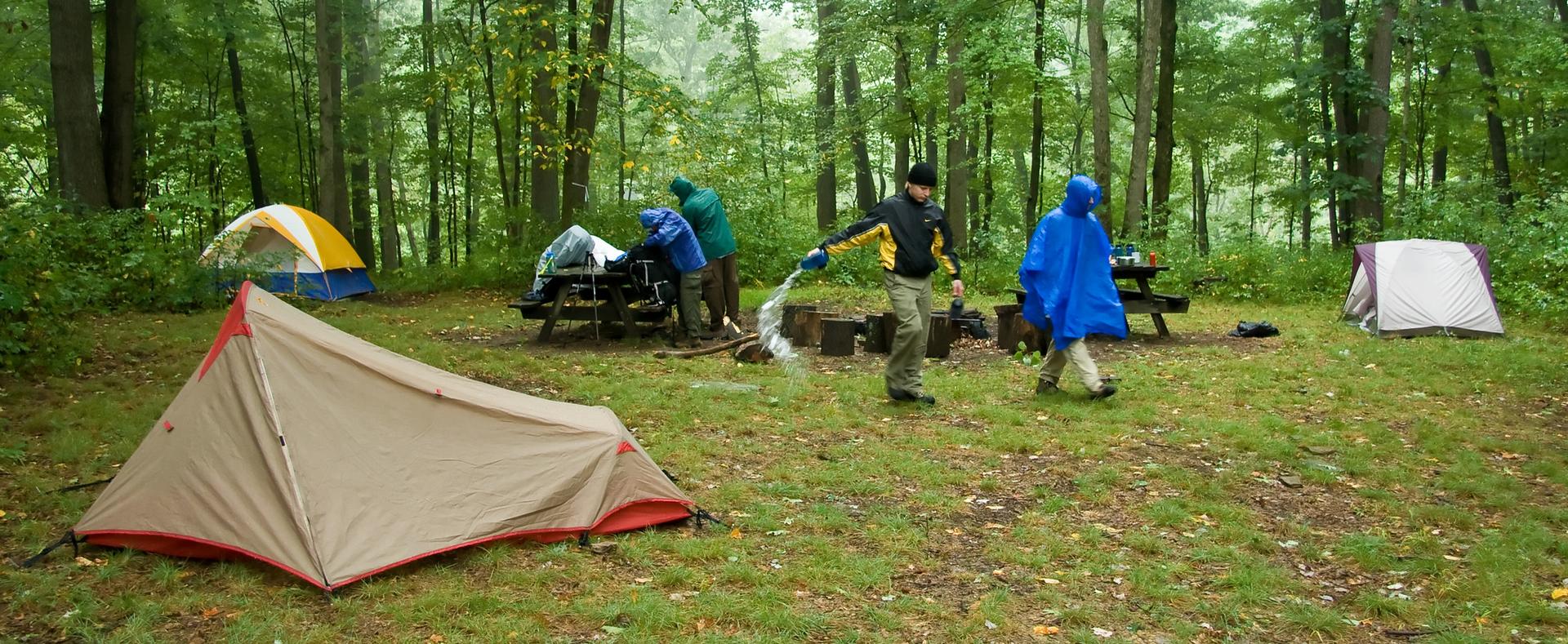 People in rain gear at a forest campsite with multiple tents on a rainy day.