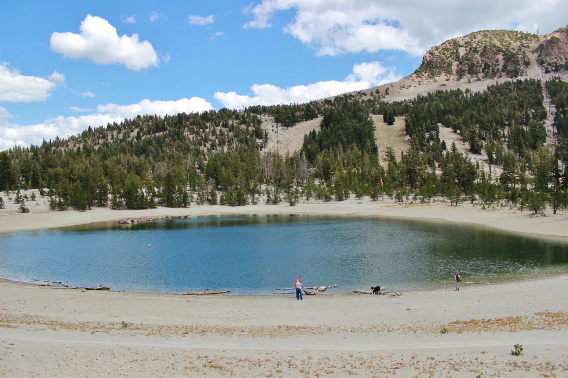 Sunny day with a blue lake, sandy shore, forested mountain, and people.
