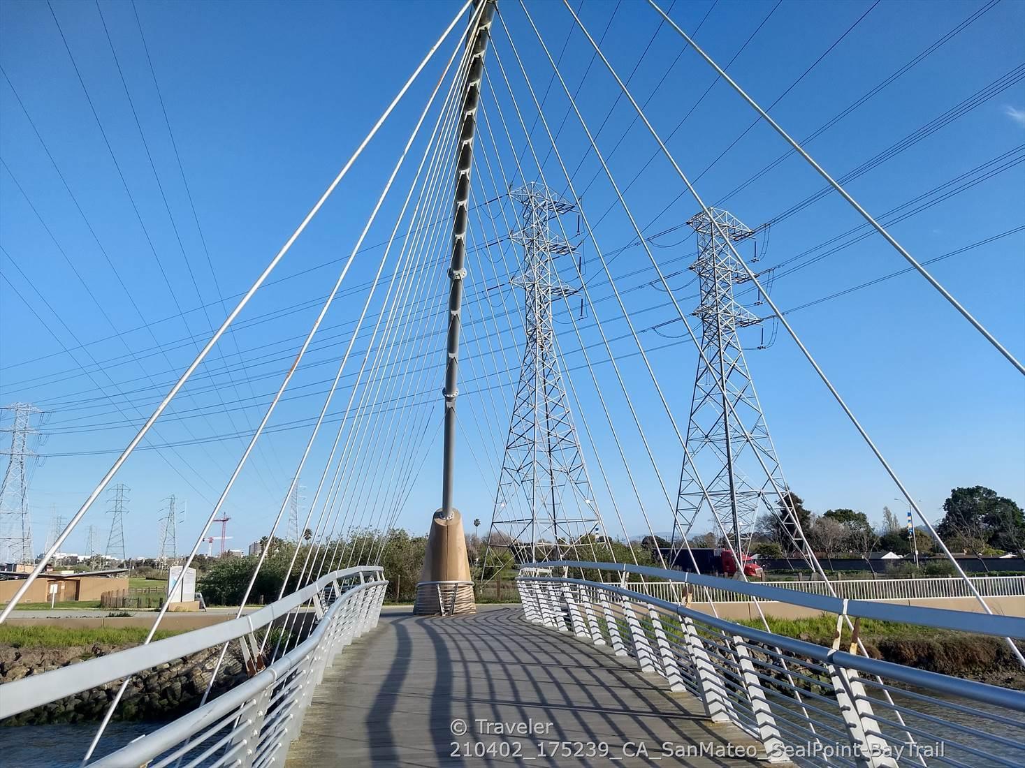 Modern pedestrian bridge with cables, power lines, and towers under a clear blue sky.
