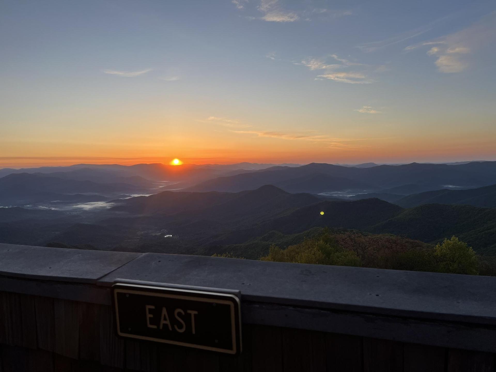 Golden sunrise over distant mountains and valleys, with an 'EAST' sign in foreground.