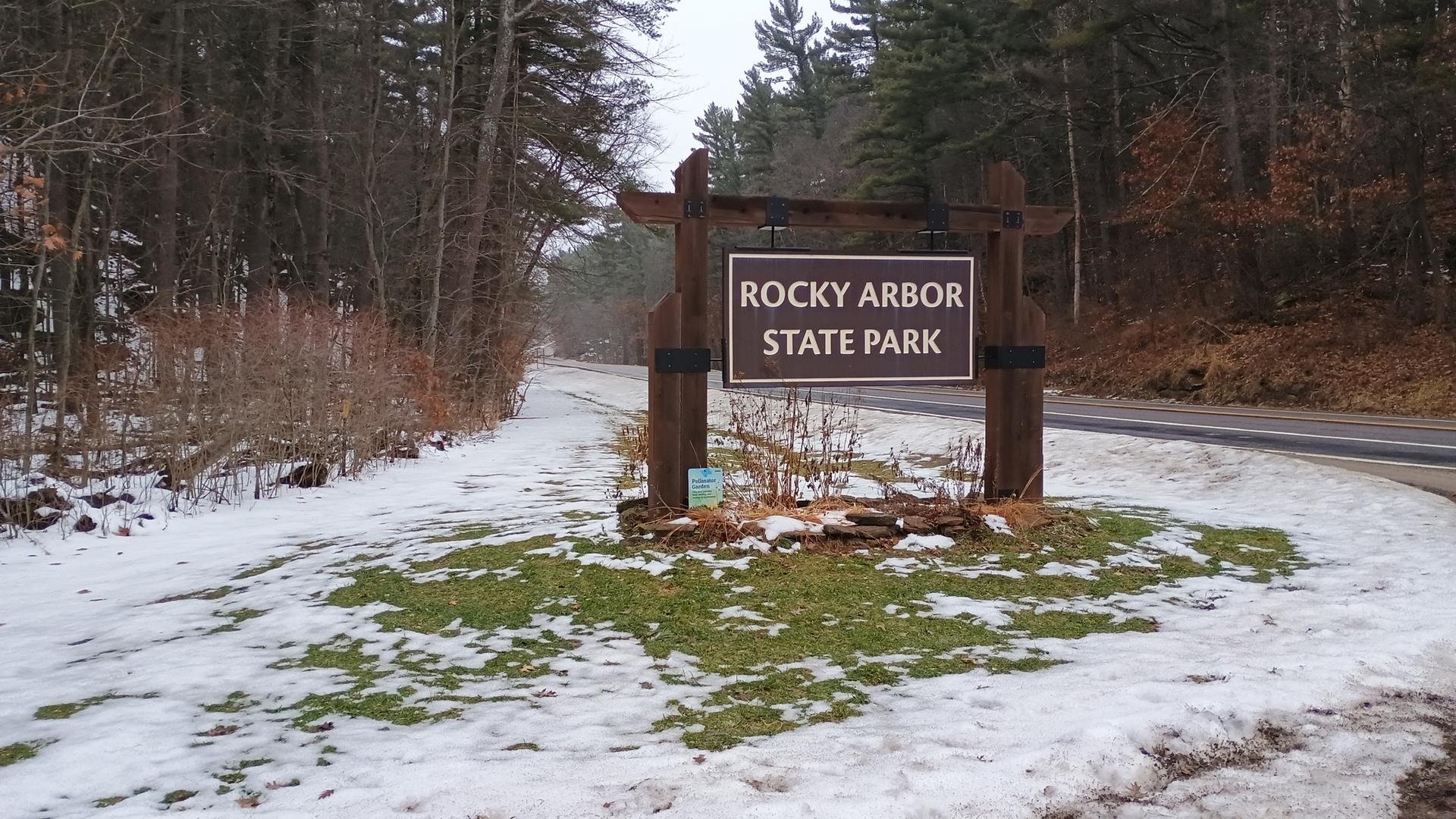 Wooden sign by a road with melting snow and bare trees under overcast sky.