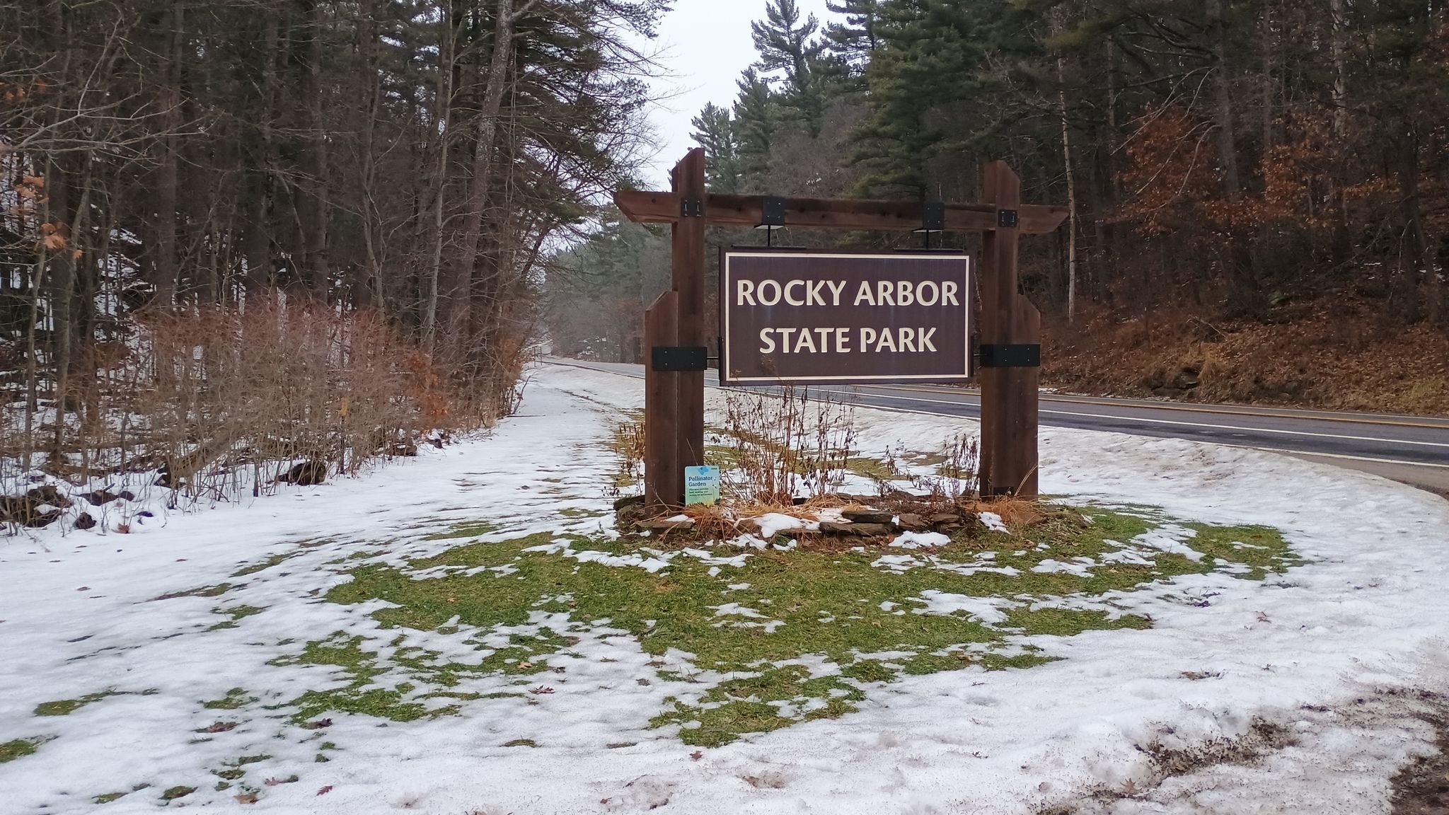 Wooden sign by a road with melting snow and bare trees under overcast sky. - Rocky Arbor Loop