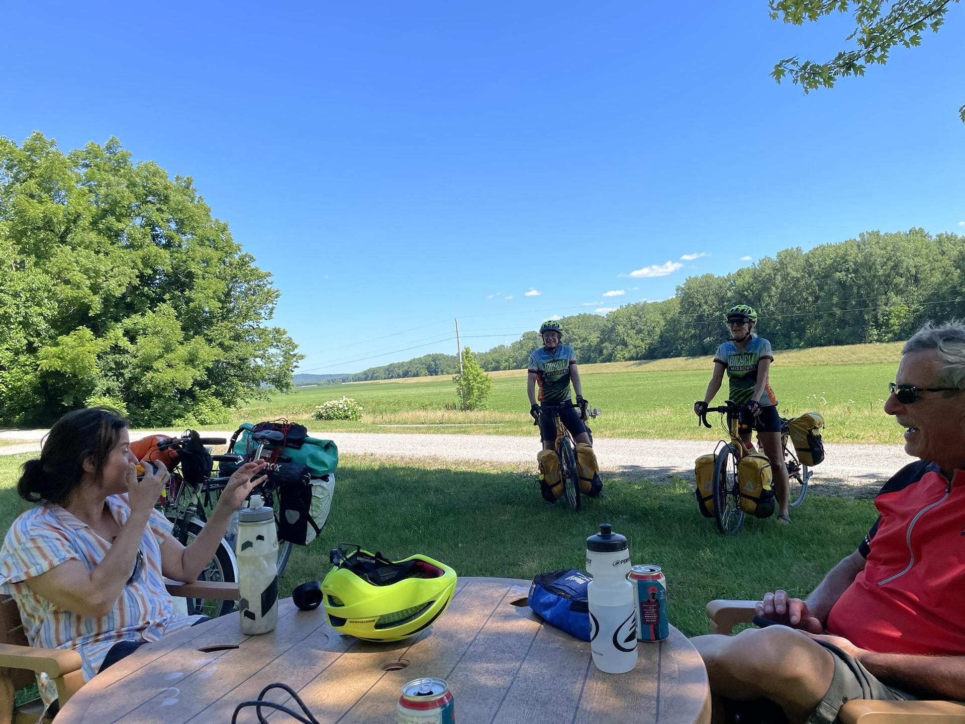 Four people relaxing outdoors, two on bikes, two at a picnic table.