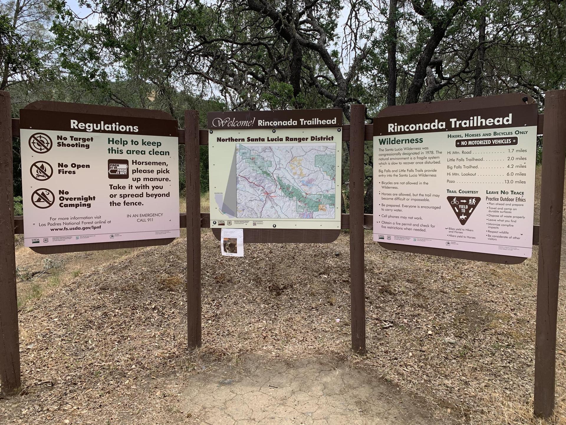 Three trailhead signs with maps and regulations on a dirt path under an overcast sky.