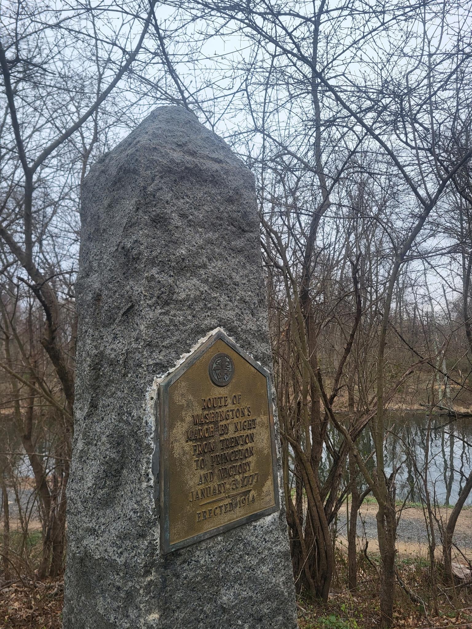 Stone monument with a bronze plaque, surrounded by bare trees and water.
