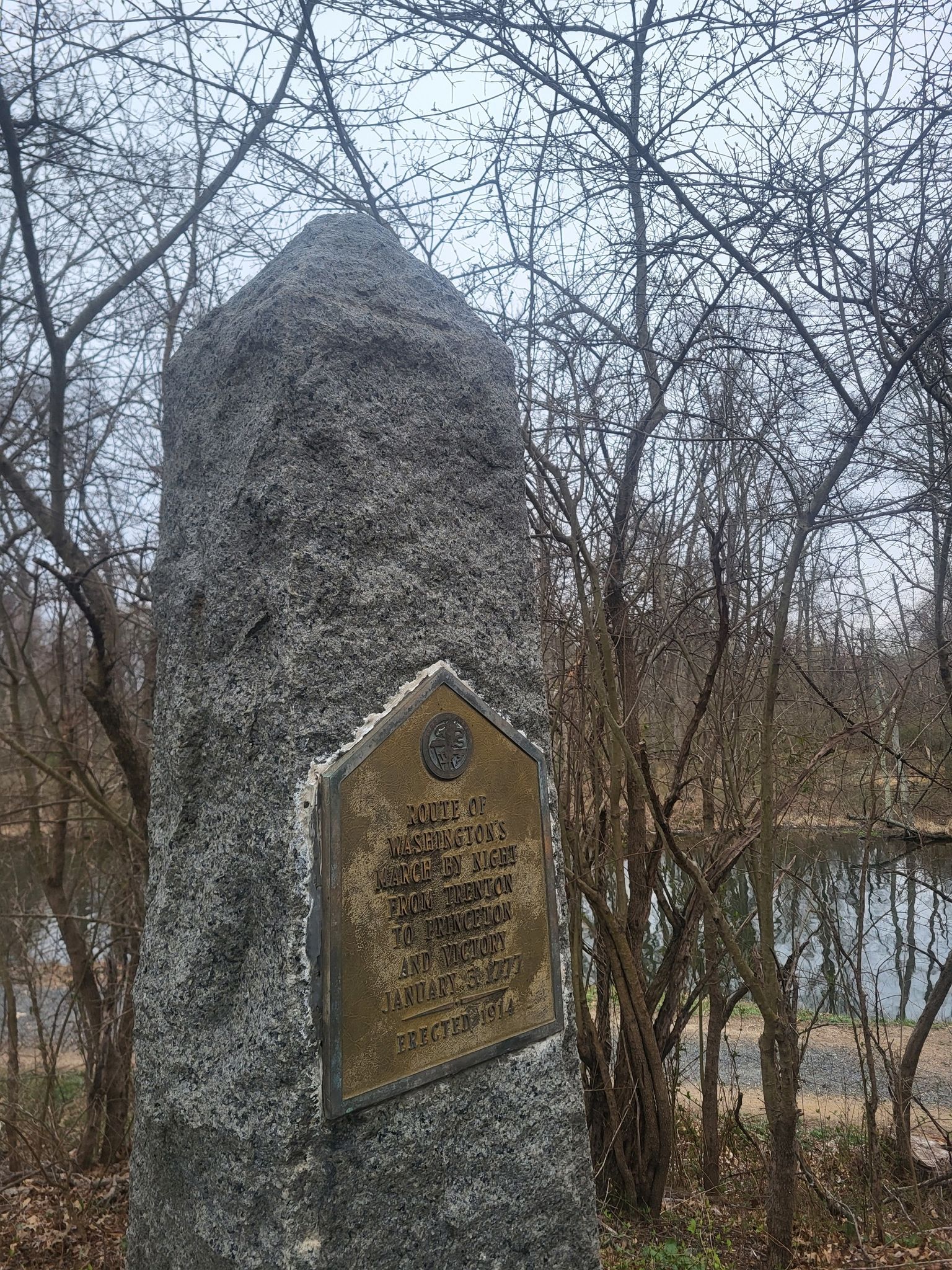 Stone monument with a bronze plaque, surrounded by bare trees and water. - D&R Canal Bridge loop from MarketFair