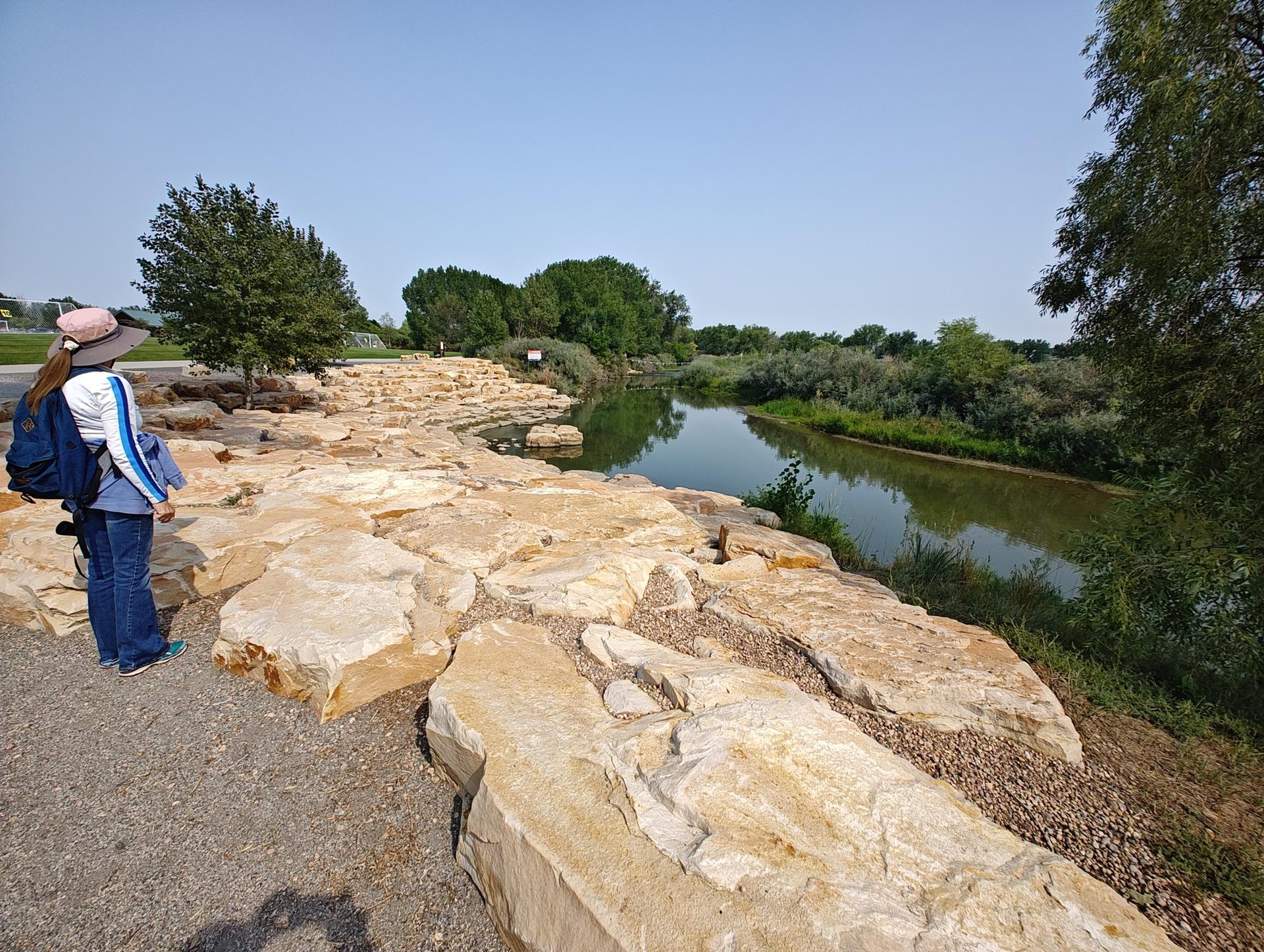 Person with backpack on rocky bank overlooking a river on a sunny day.