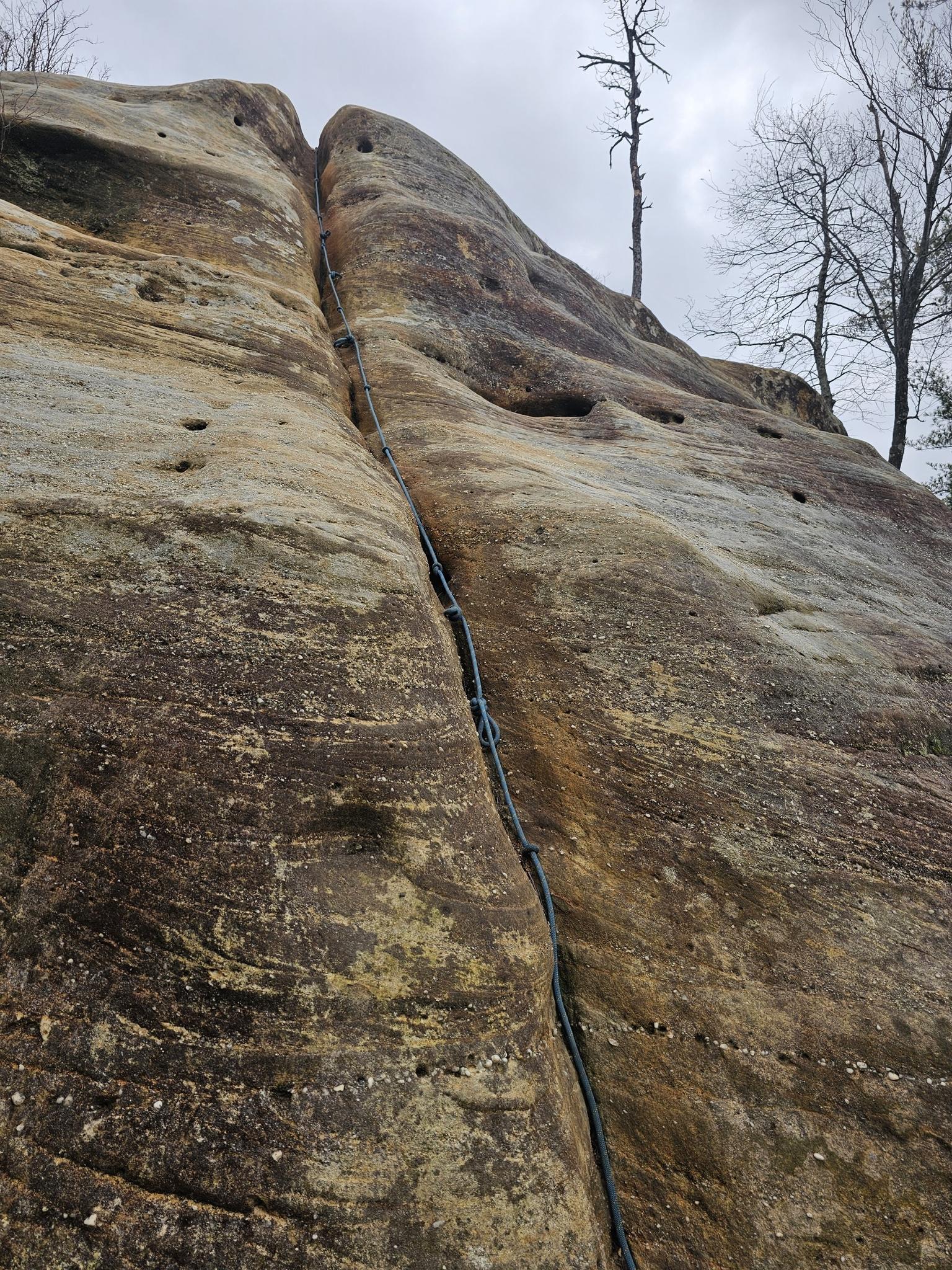 A climbing rope ascends a large, textured rock face under an overcast sky.