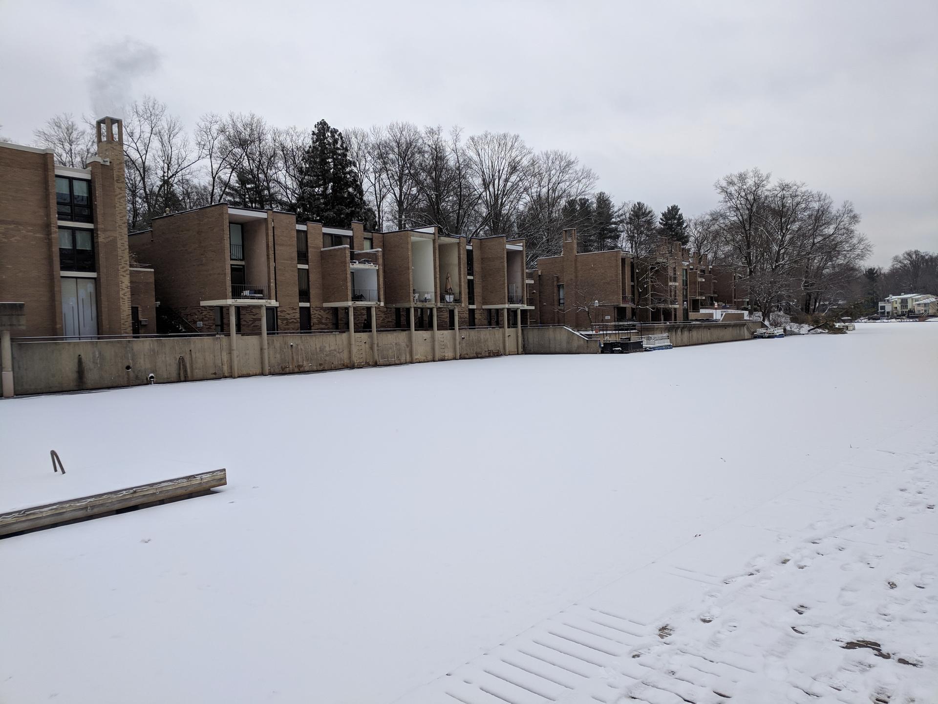 Snow covers a frozen lake and modern brick buildings under a gray sky.
