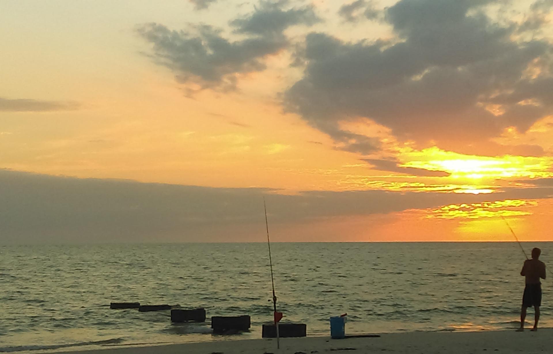 A person fishes in the ocean during a vibrant sunset from a sandy beach.