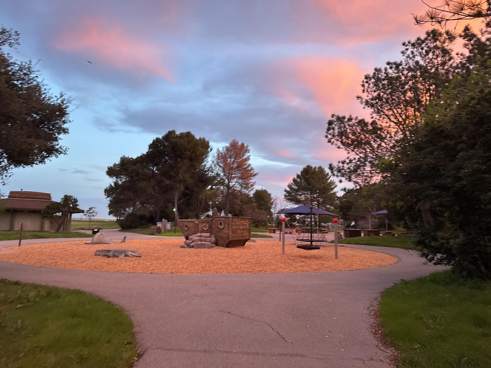 Playground with wooden structures and wood chips under a colorful sunset sky. - Promenade Trail and Bluff Loop
