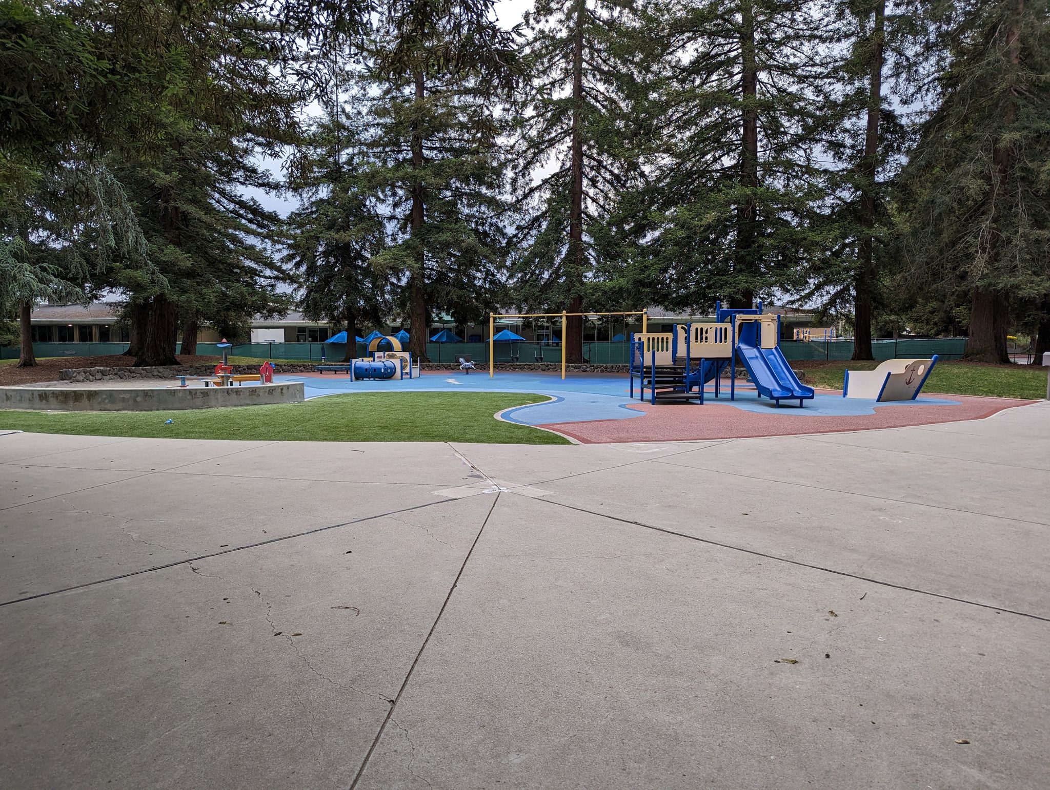 Empty playground with blue equipment, tall trees, and concrete path under an overcast sky. - Central Park loop