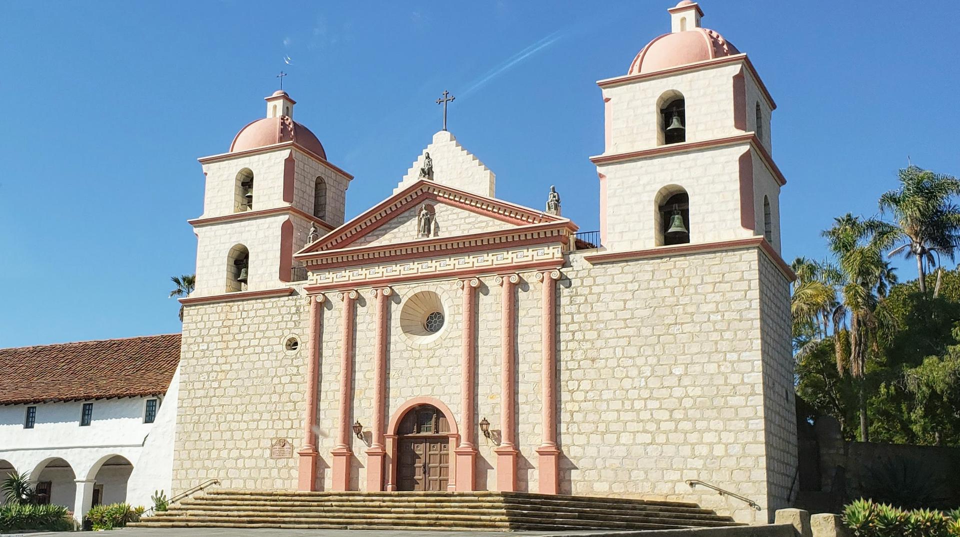 Ornate stone church with pink domes and bell towers under a blue sky.