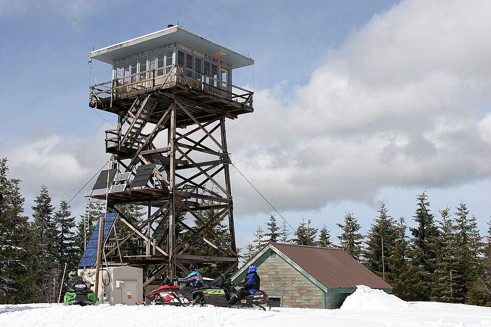 A tall wooden fire lookout tower, snowmobiles, and a person in a snowy forest.