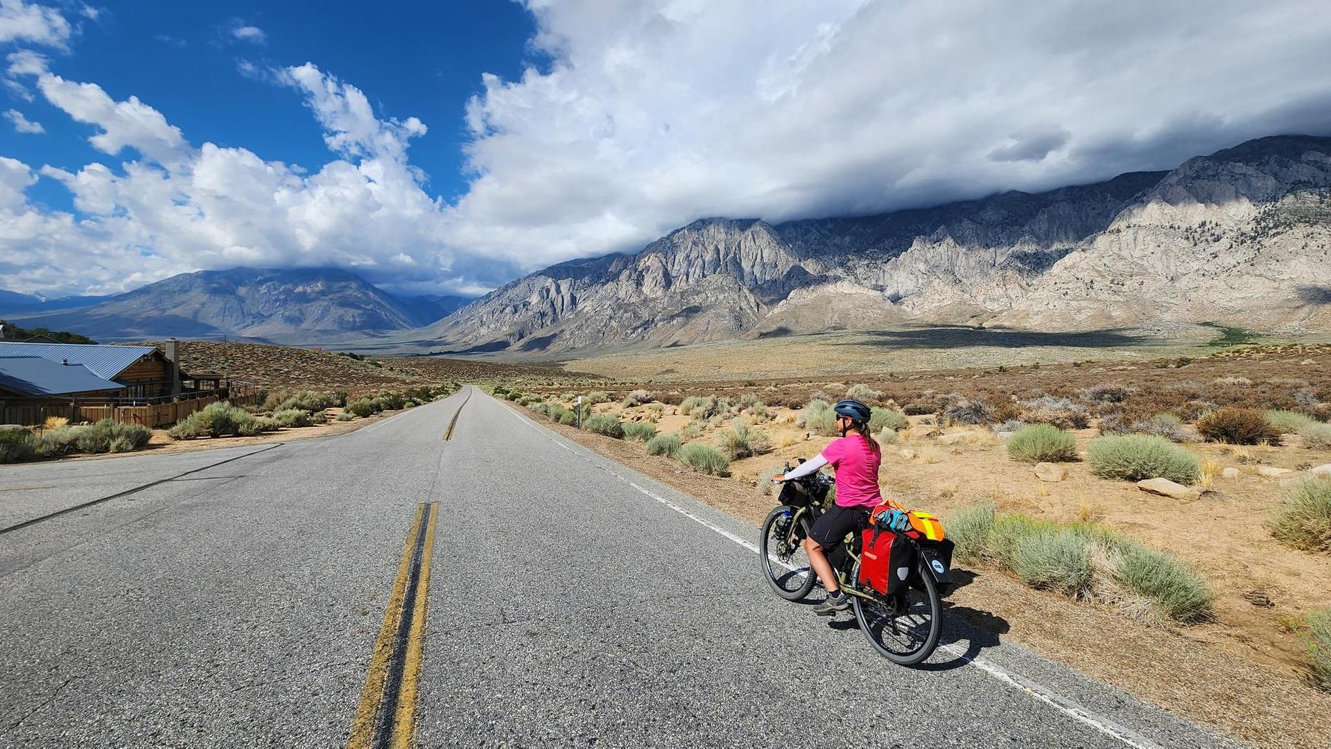 Cyclist with gear stopped on a long road in an arid mountain landscape.