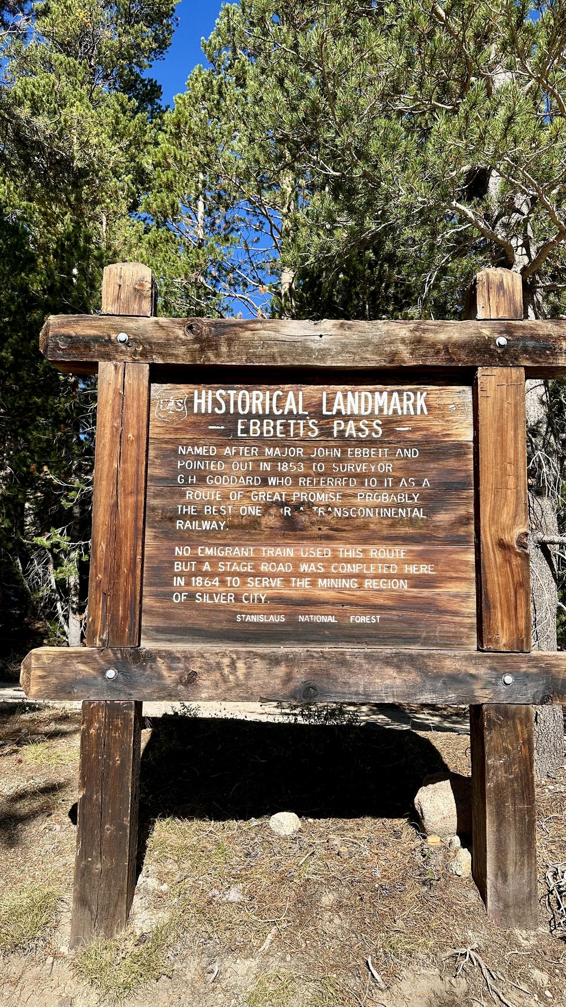 A large wooden historical landmark sign surrounded by tall pine trees.