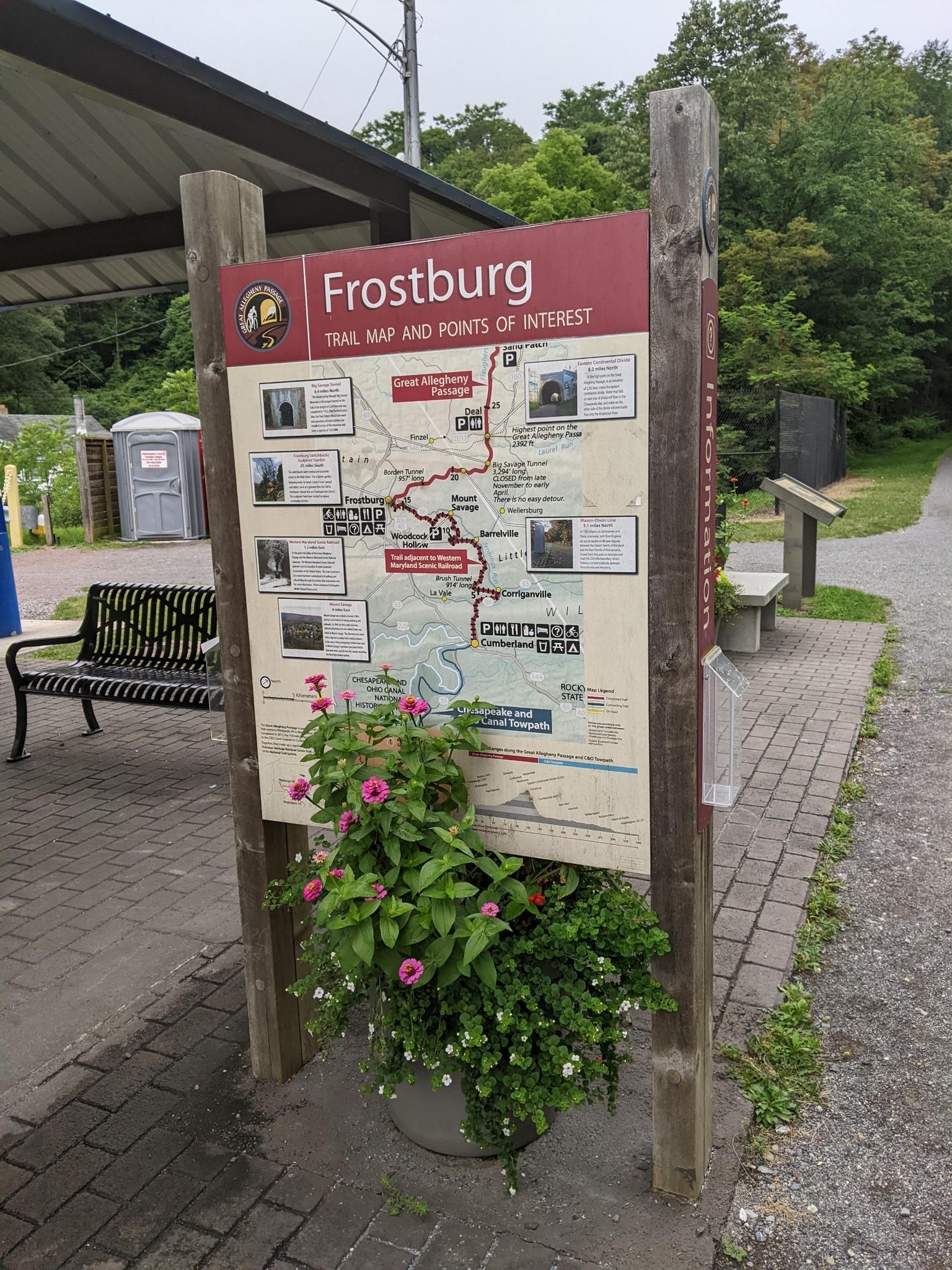 An outdoor trail map sign with flowers, benches, and a portable toilet.