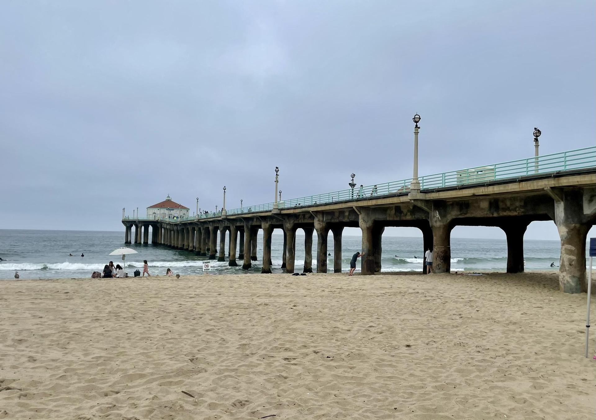 A long pier extends over a sandy beach and ocean under a cloudy sky.