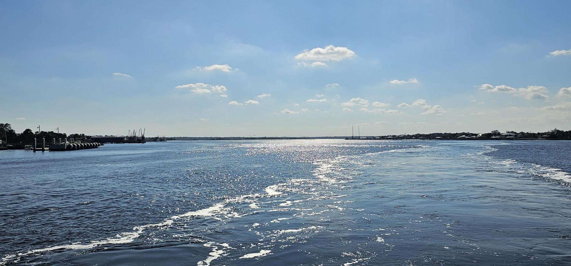 Wide river with boat wake, distant boats, and sun glare on the water.