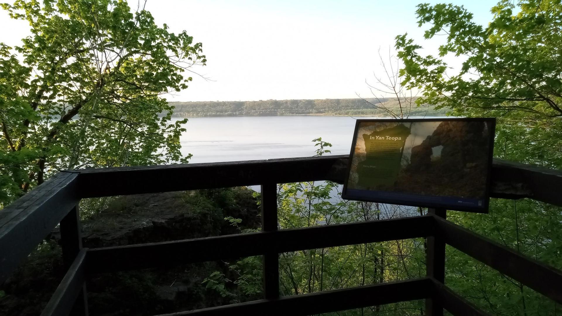 Wooden observation deck with an interpretive sign overlooking a sunny lake.