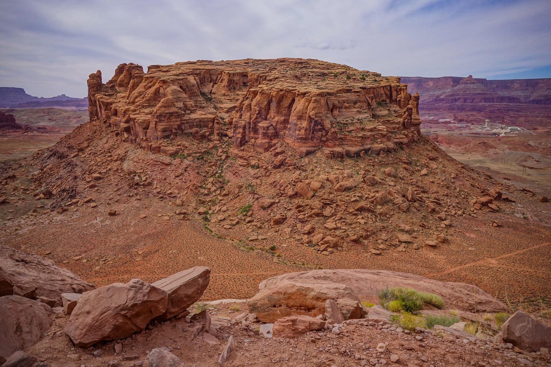 Large red rock mesa dominates an arid, rocky desert landscape under a cloudy sky.