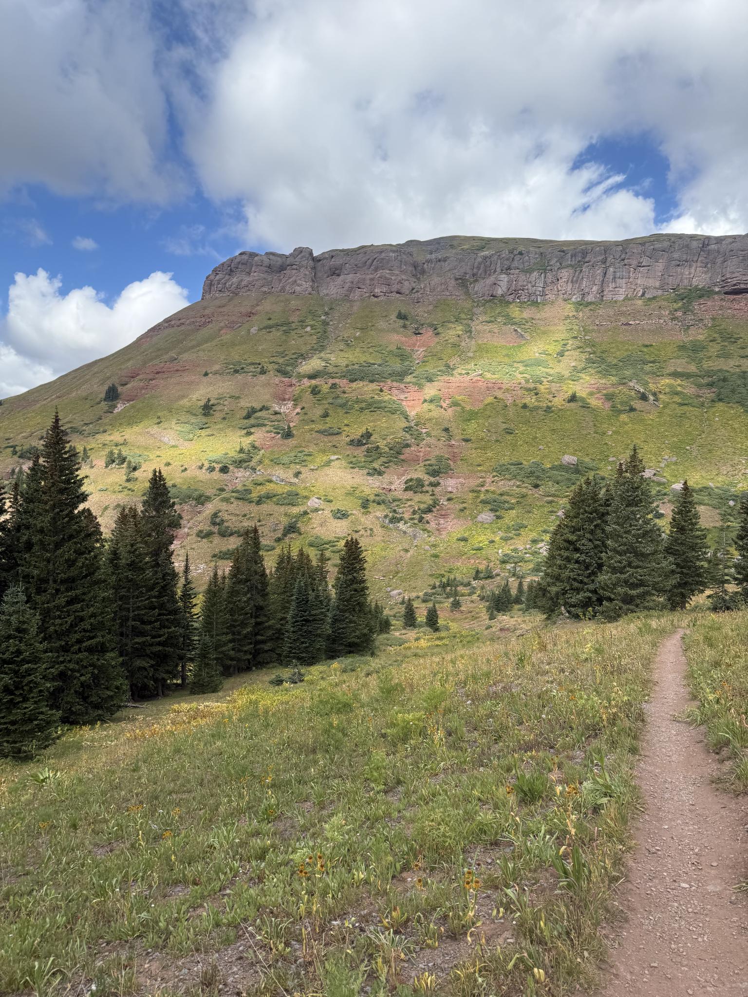 A dirt trail leads towards a large, green mountain under a cloudy sky.