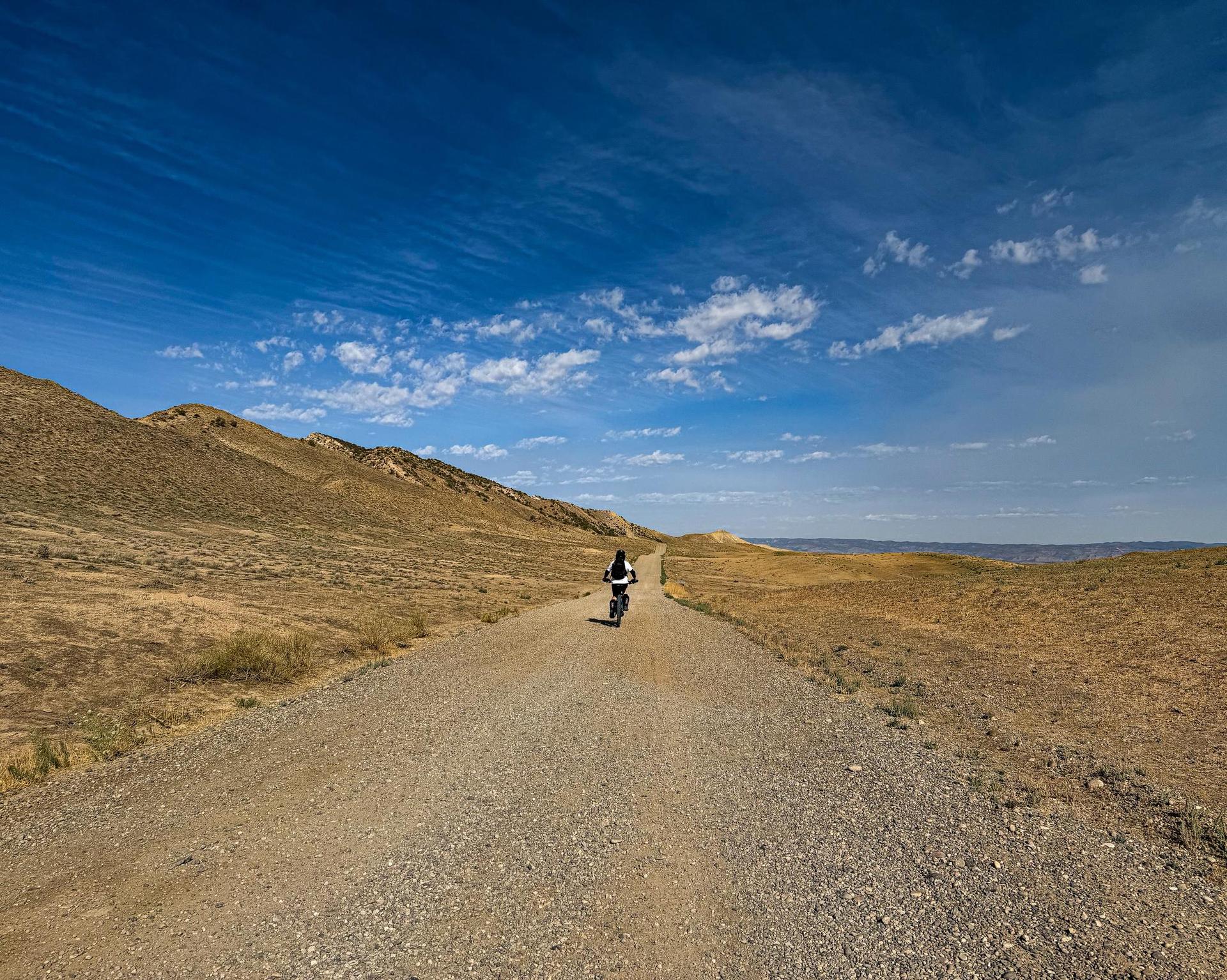 Dirt biker rides on a long, straight dirt road under a vast blue sky.