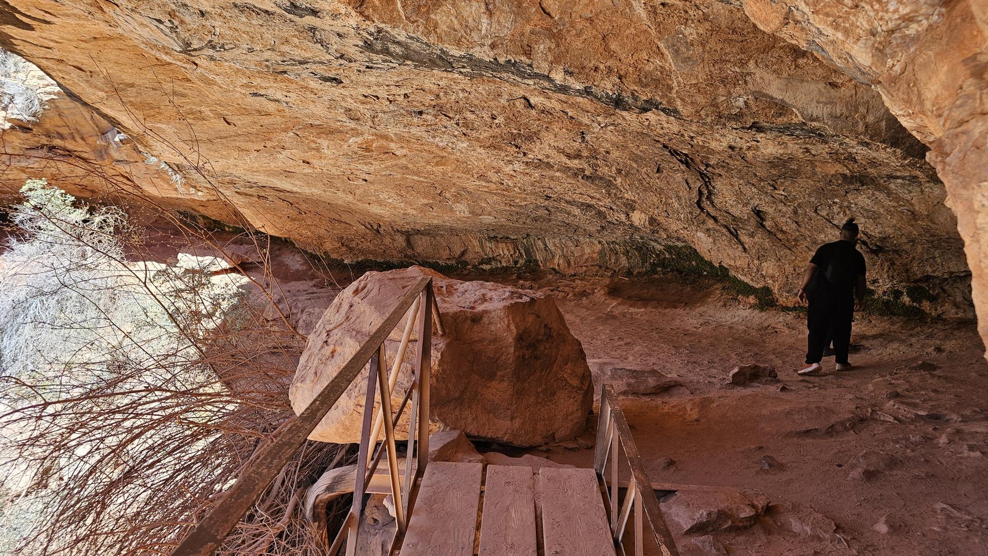 A person stands on a wooden walkway under a large, dark rock overhang.