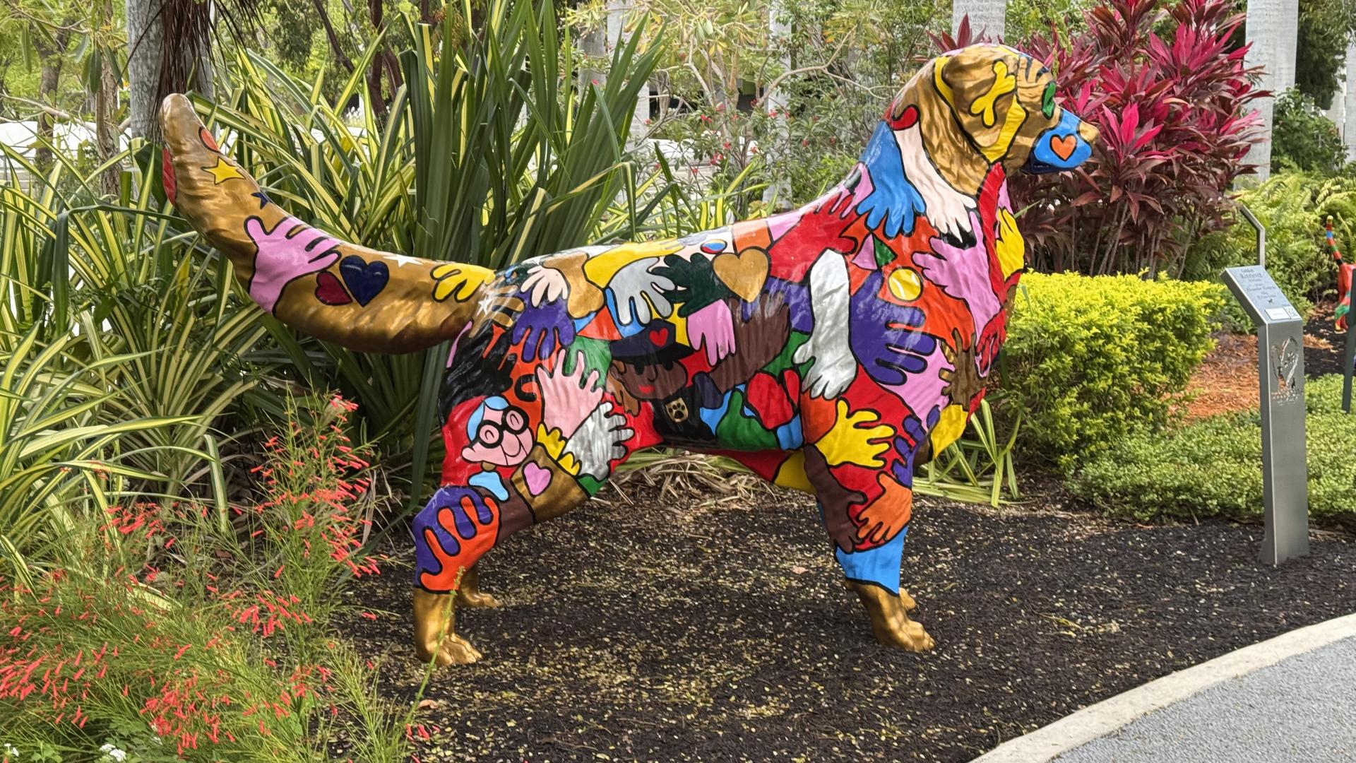A colorful dog statue, painted with hands, stands in a green garden.