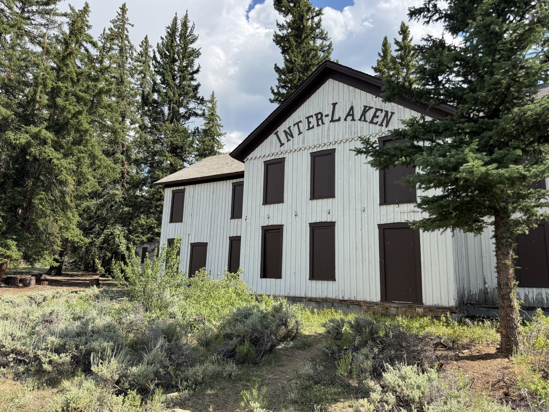 Large white historic building with dark windows, surrounded by pine trees.