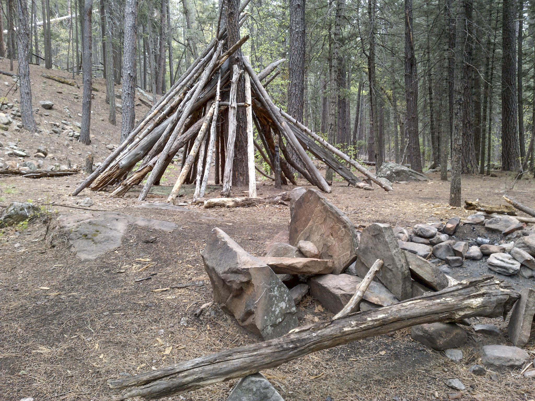 A log teepee structure and stone fire pit in a forest clearing.
