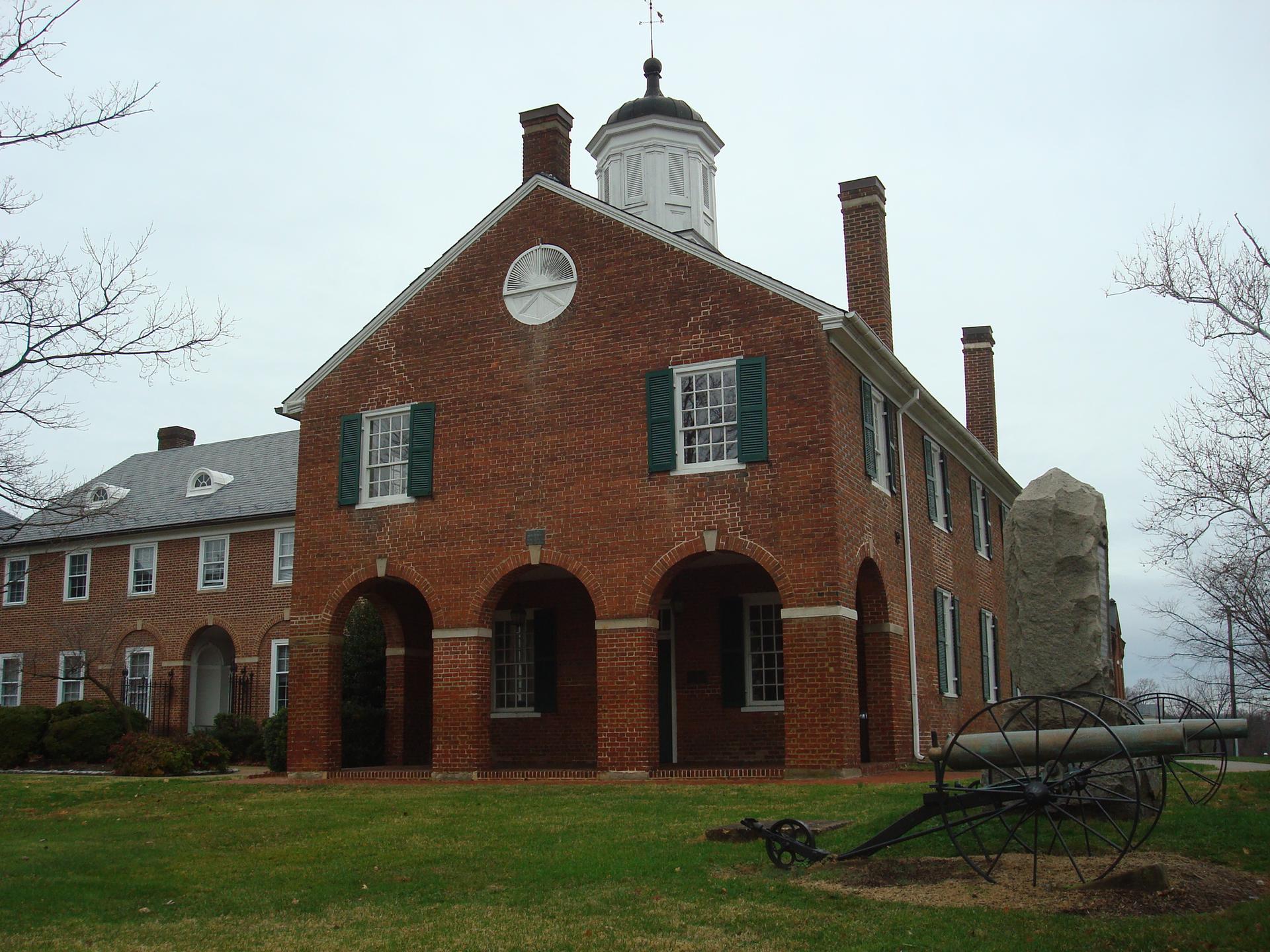 Brick building with cupola, arches, and cannon on a grassy lawn under an overcast sky.