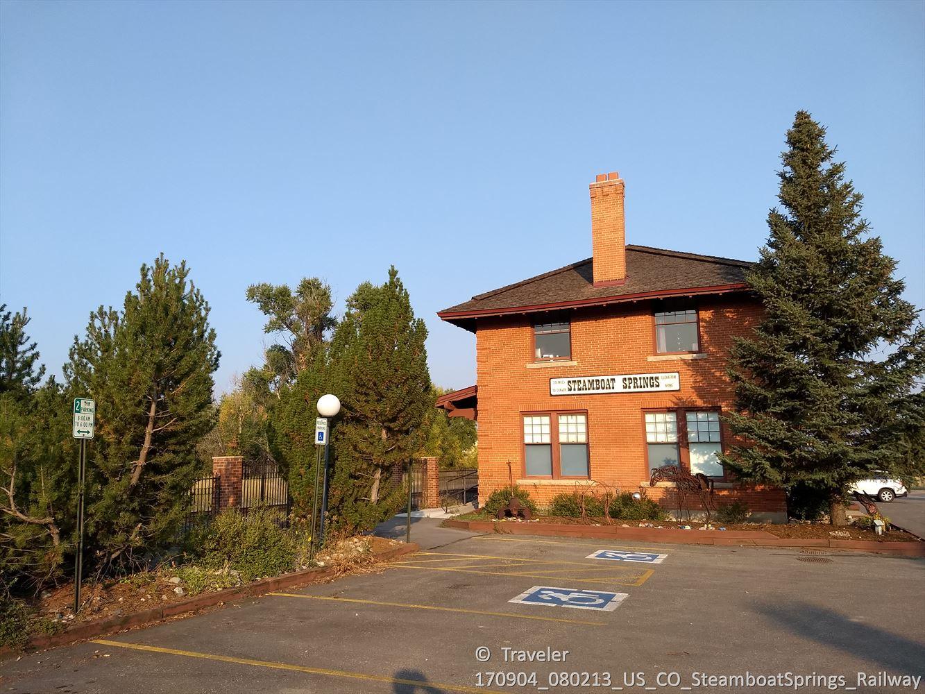 Brick building with a parking lot and tall pine trees under clear sky.