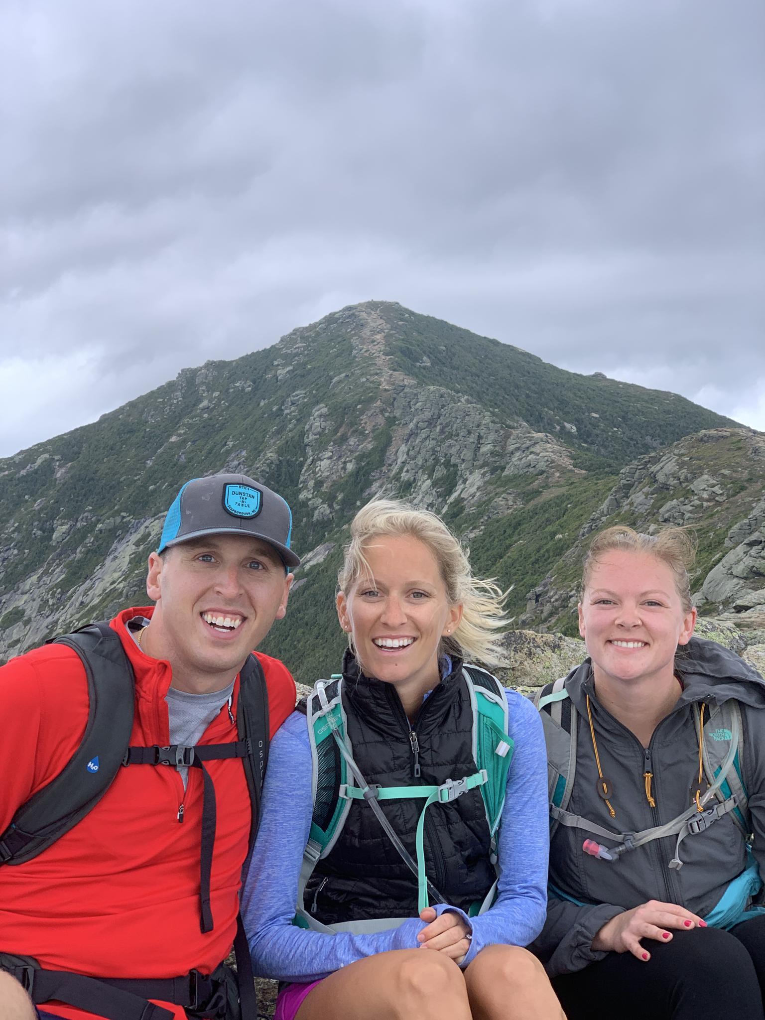 Three smiling hikers on a mountain summit with a peak behind them.