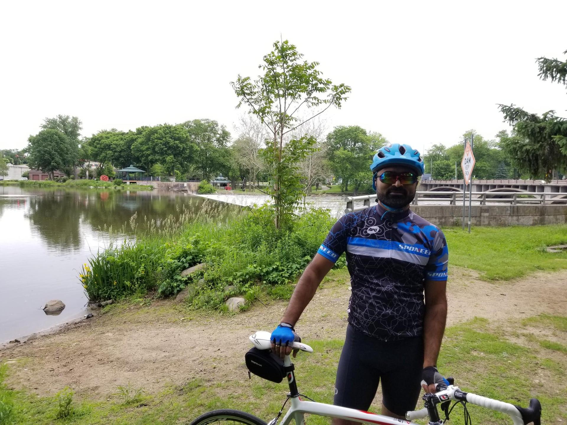 Cyclist in blue gear stands with bike by a lake on an overcast day.