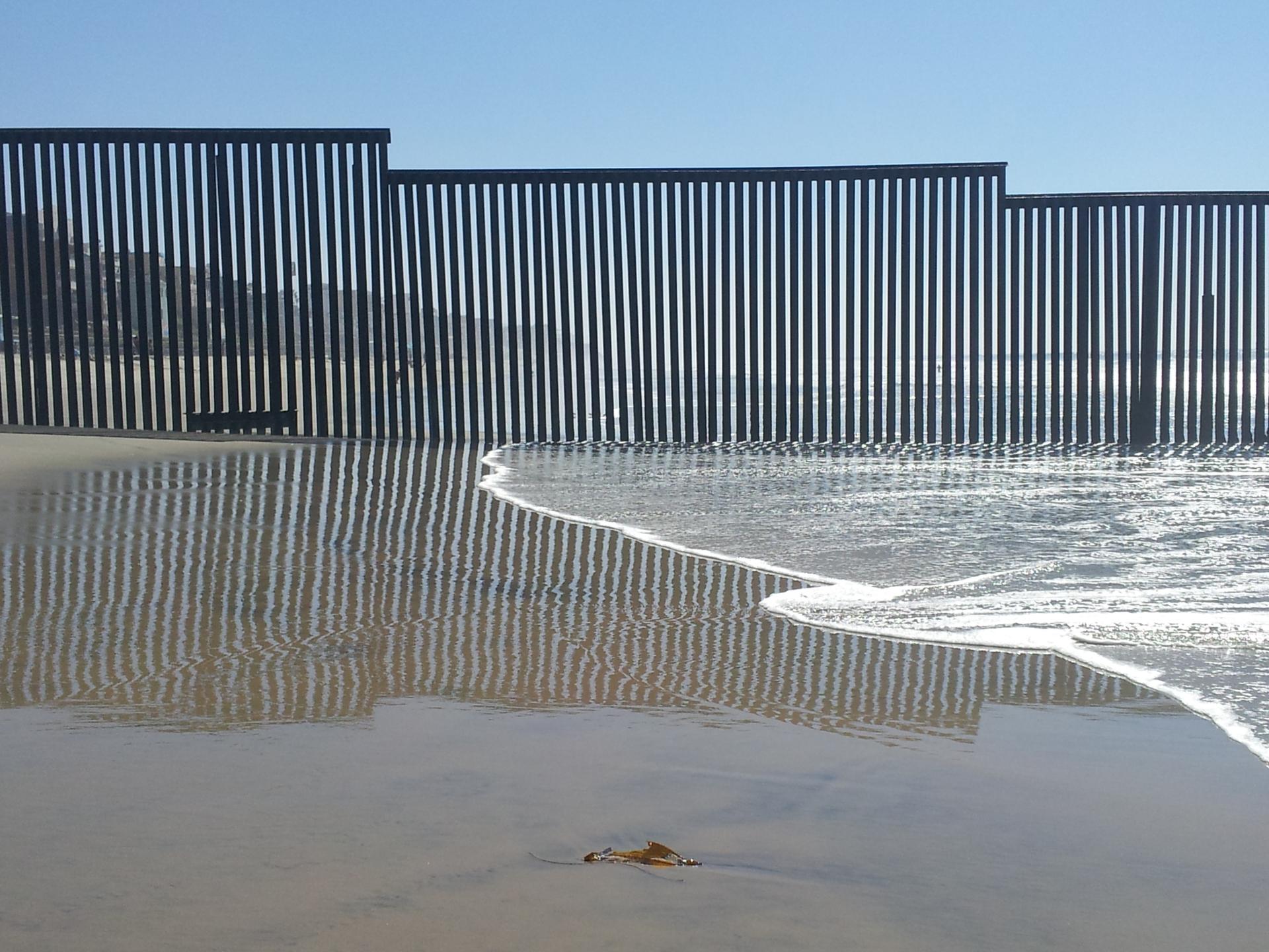 Sunlight reflects off wet sand and a dark fence at the ocean's edge.