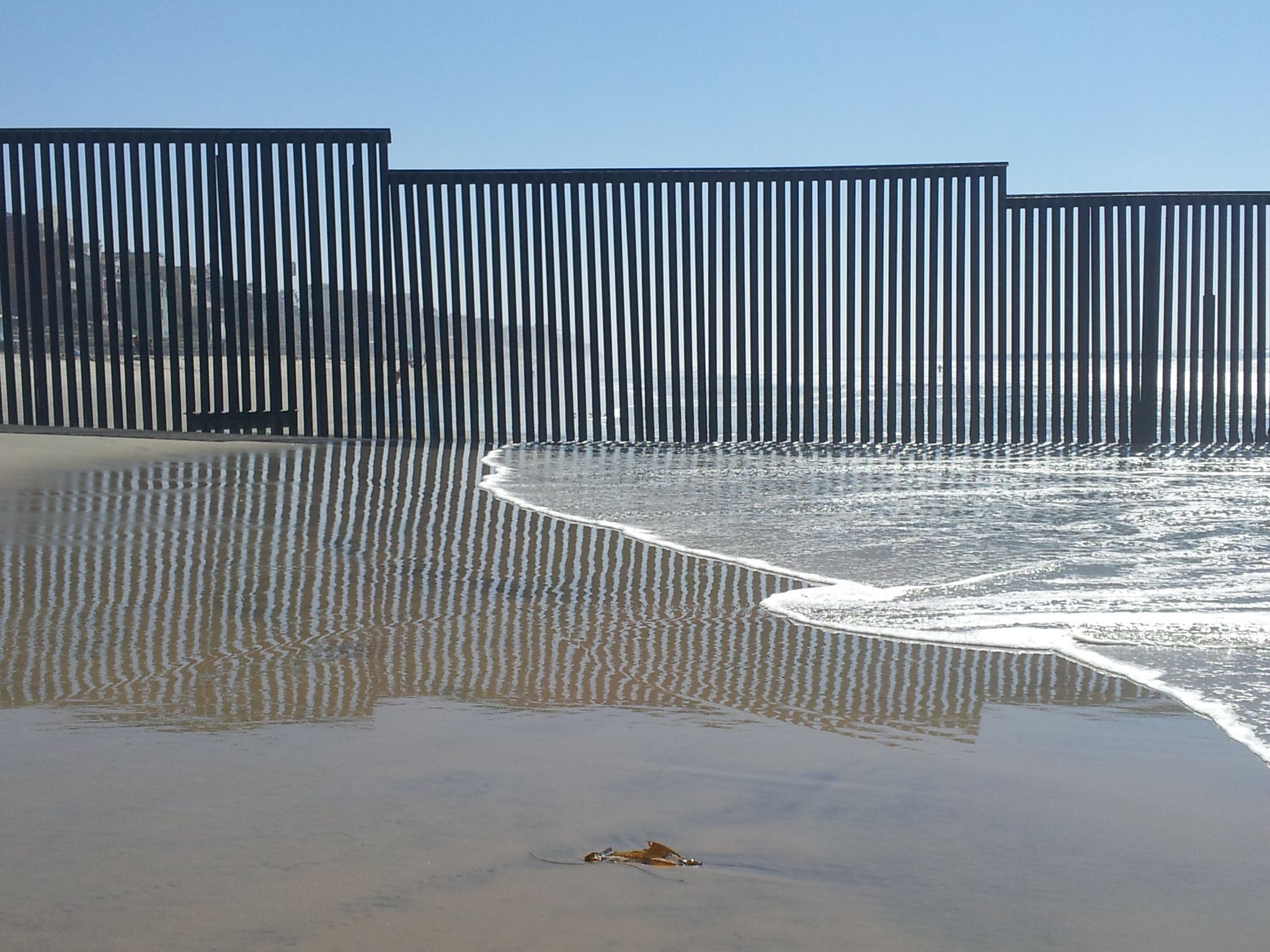 Sunlight reflects off wet sand and a dark fence at the ocean's edge. - Paseo Costero Trail