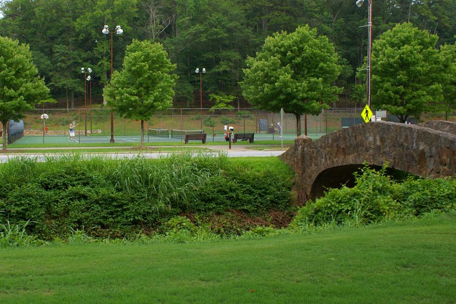 Stone bridge overlooks green park with people playing tennis under bright sky.